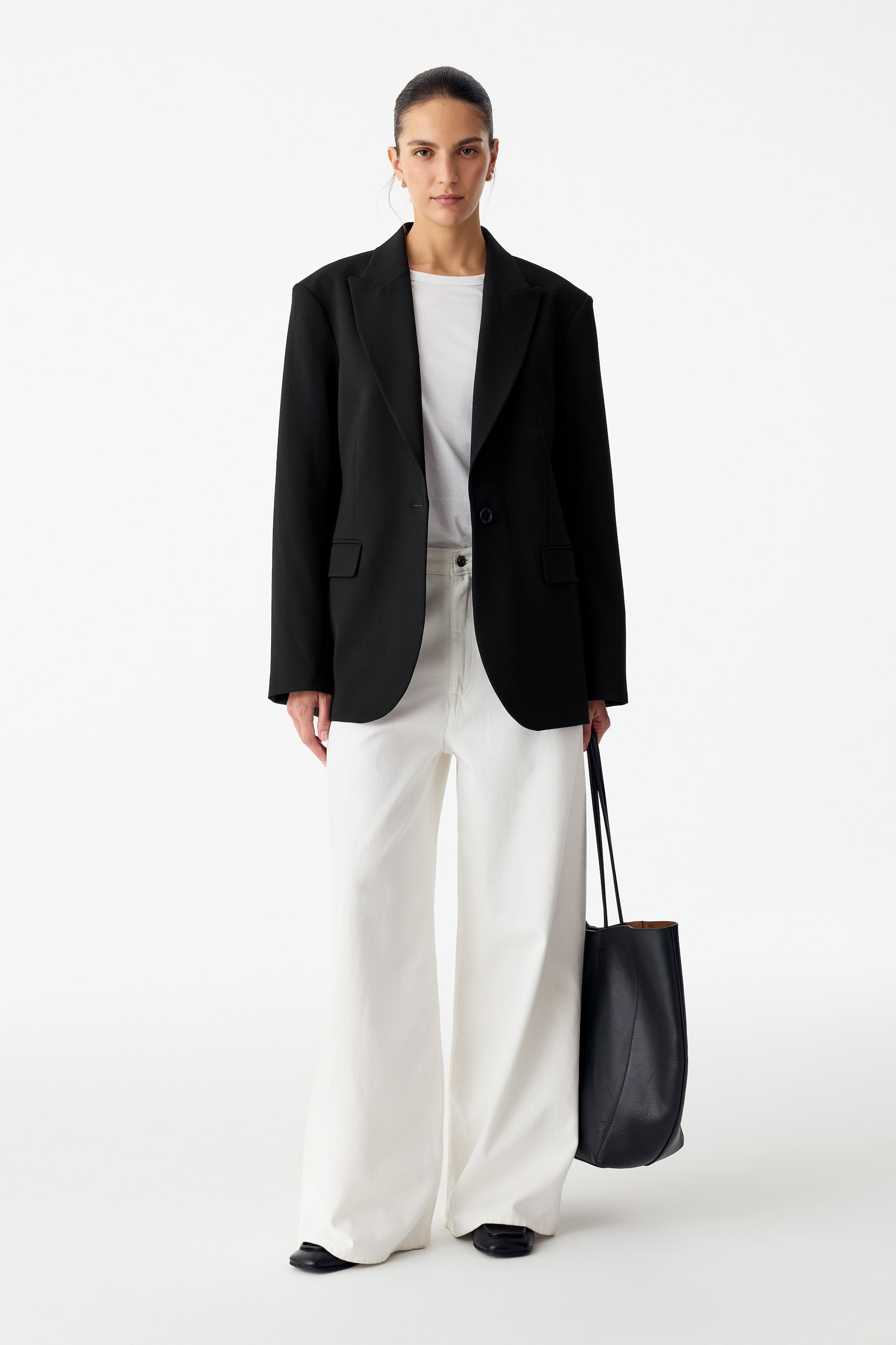 Größeres Bild anzeigen: Ein schwarzer, einreihiger Blazer mit Reverskragen und Pattentaschen wird vom Model offen über einem weißen T-Shirt und weiten weißen Hosen getragen. Das Model trägt außerdem eine schwarze Tote-Bag aus Leder.