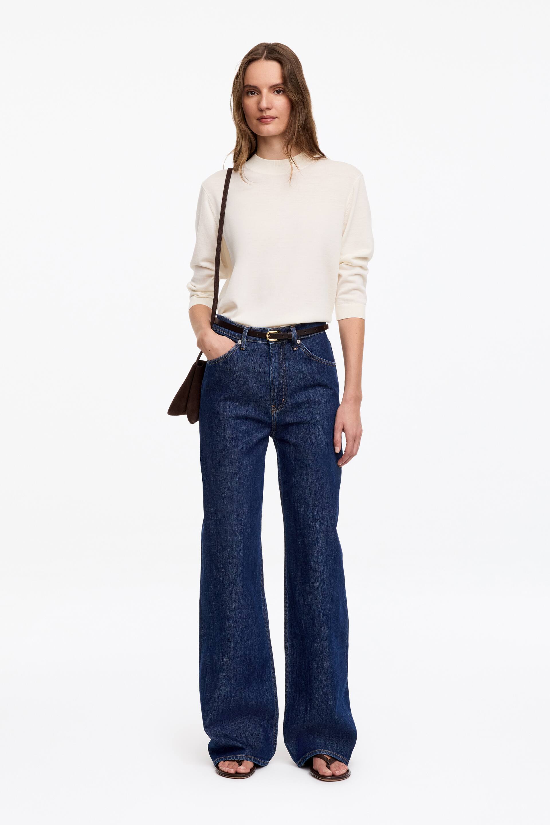 Agrandir l'image: Les jeans LUPINE présentent un lavage bleu foncé, une taille haute et une coupe ajustée qui s'évase en jambes larges. Le mannequin porte les jeans, qui sont assortis à un haut de couleur claire rentré à l'intérieur, et une ceinture foncée est visible à travers les passants de ceinture. Une des mains du mannequin repose dans une poche avant.