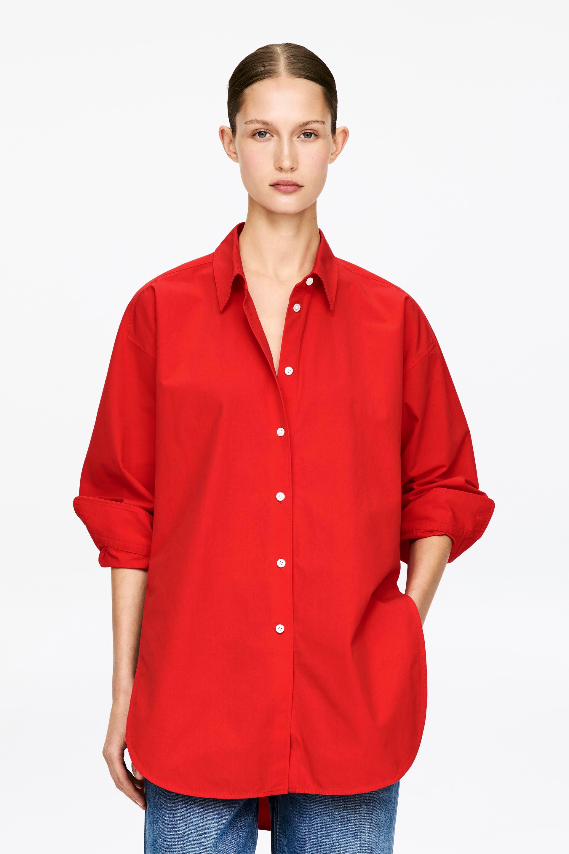 Größeres Bild ansehen: Ein rotes Hemd mit Knopfleiste wird präsentiert, nach vorne gerichtet, mit aufgerollten langen Ärmeln auf Dreiviertellänge. Es zeichnet sich durch einen spitzen Kragen, weiße Knöpfe vorne und eine lockere Passform aus, mit einer Hand, die in einer der Seitentaschen ruht und einem abgerundeten Saum, der über blauen Jeans sichtbar ist.