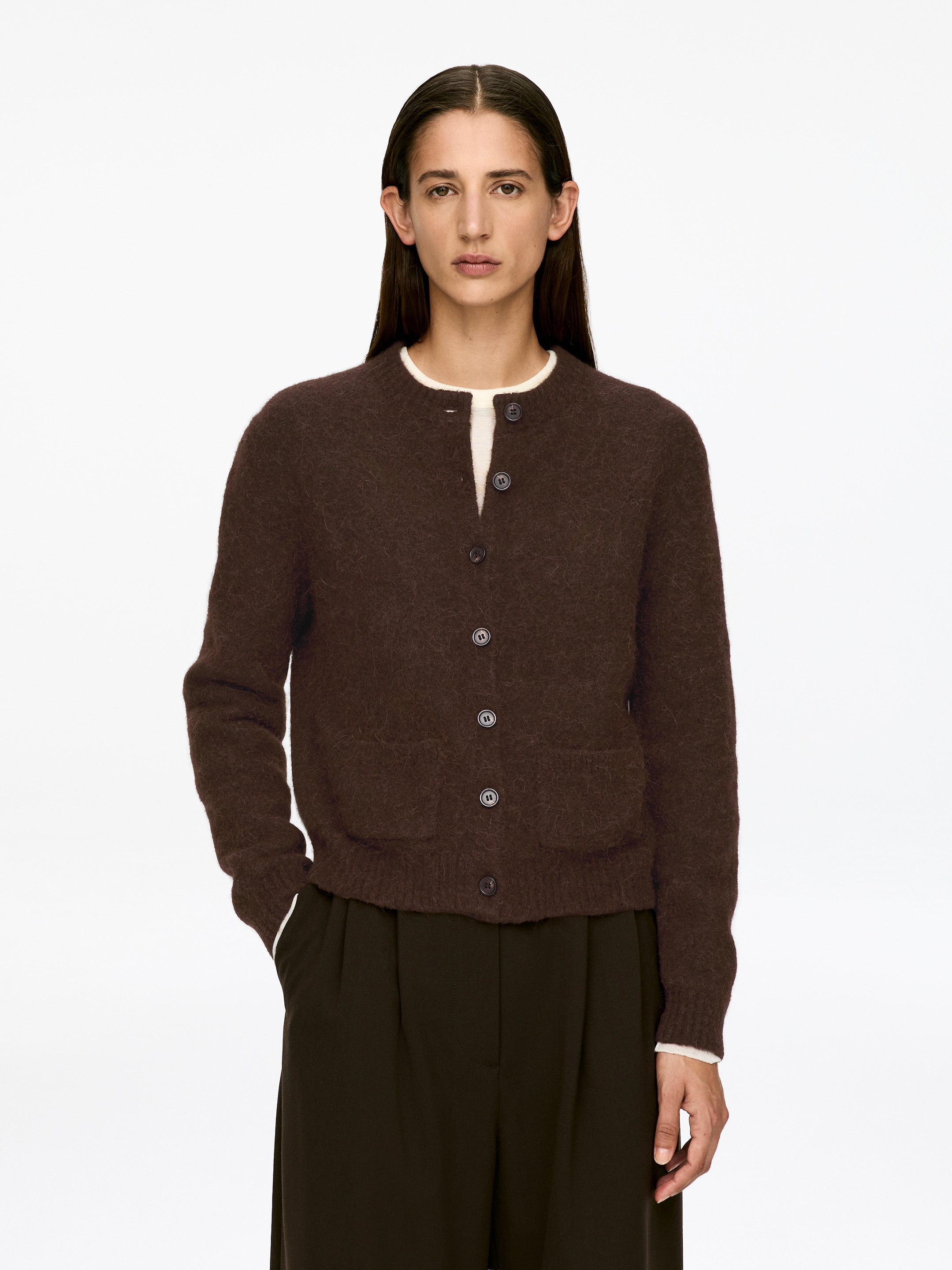 Agrandir l'image: Un cardigan tricoté marron foncé, avec un col rond, une fermeture à boutons sur le devant et deux poches plaquées, est porté par le mannequin. Le cardigan a une texture douce et légèrement duveteuse et est superposé à un haut de couleur claire, le mannequin mettant une main dans une poche.