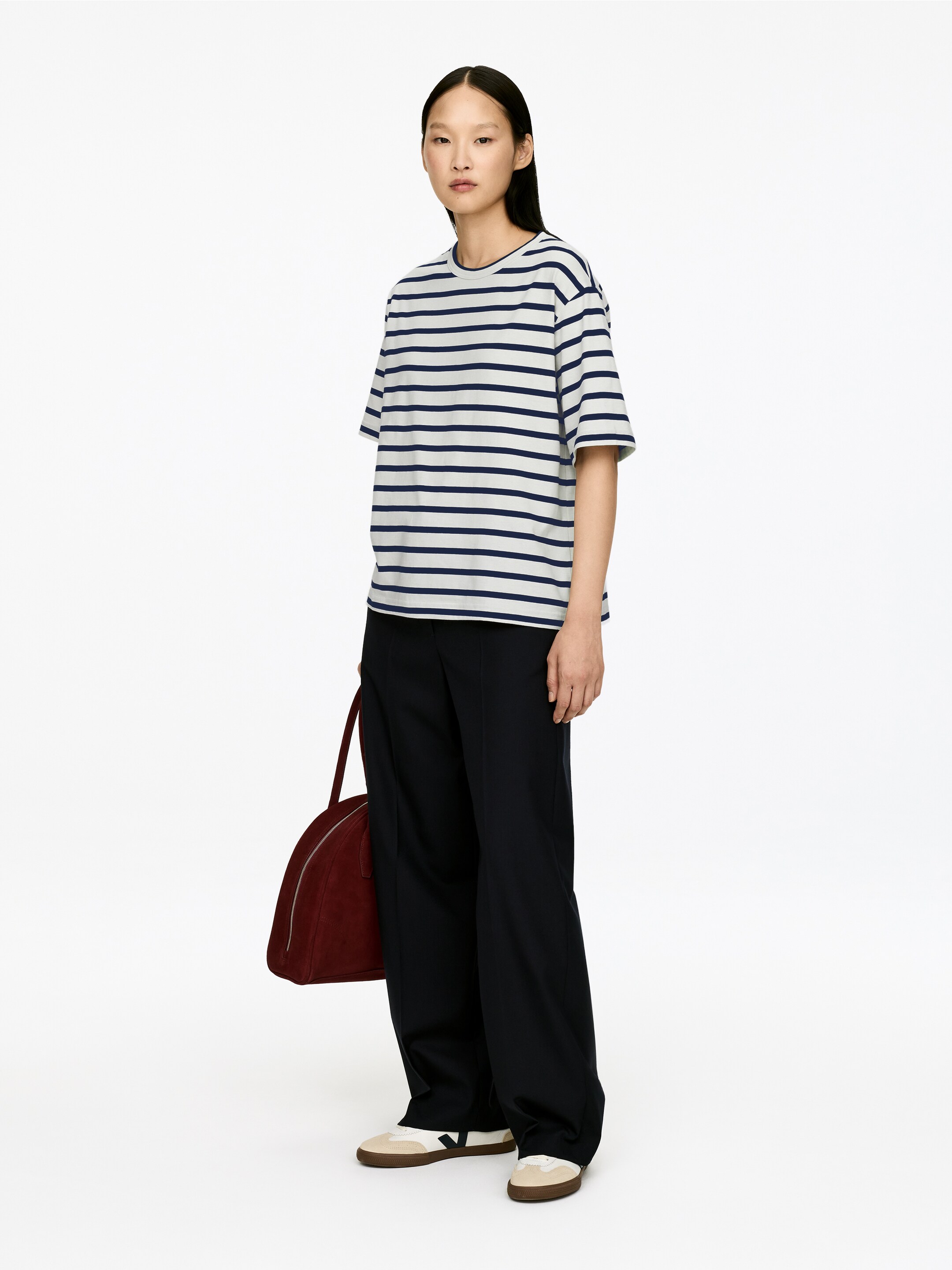 Agrandir l'image: Un t-shirt blanc à rayures horizontales bleu marine, avec un col rond et des manches courtes, est porté par une personne se tenant légèrement inclinée vers la gauche. Ils portent également un pantalon large bleu marine foncé et des baskets crème et marron, et tiennent un sac à main arrondi rouge foncé à leur côté.