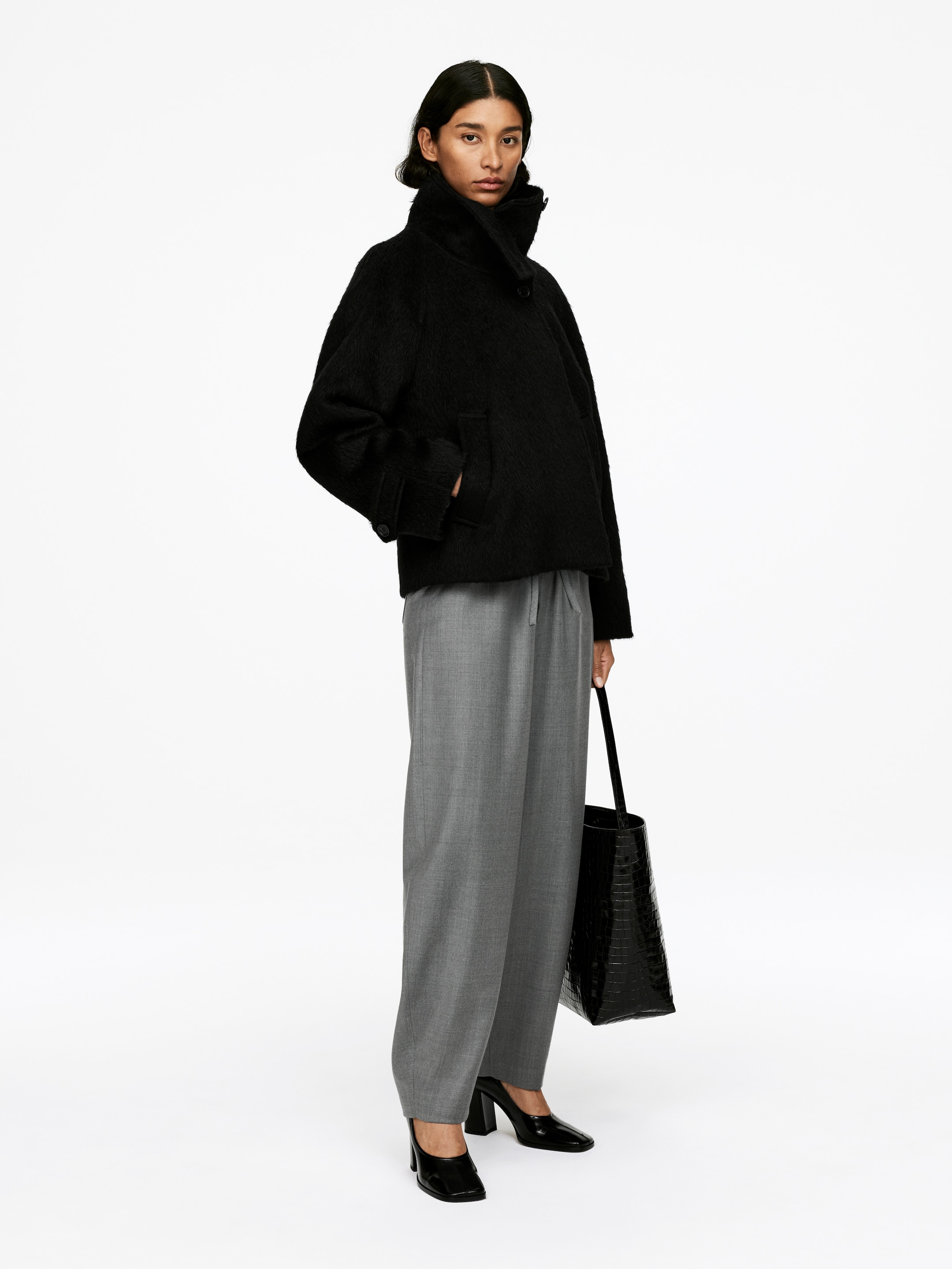Agrandir l'image: Une veste noire à texture duveteuse est présentée en vue 3/4, avec un col montant et des poches plaquées à l'avant. Elle est associée à un pantalon large gris à coupe ample, avec une taille à cordon visible. Des chaussures à talons carrés noirs et un sac fourre-tout embossé façon crocodile noir sont également portés.