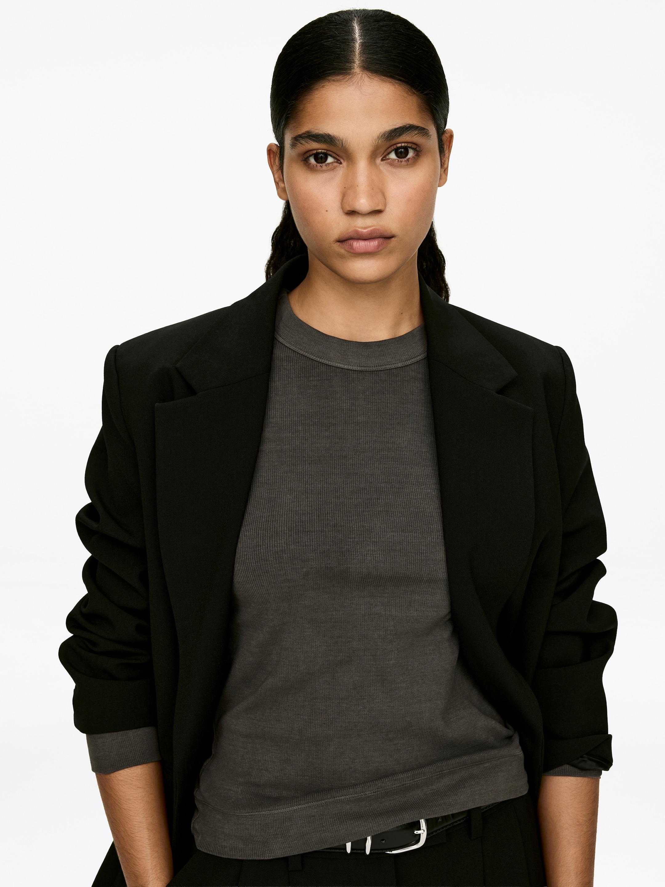 Grotere afbeelding bekijken: Een donkergrijze, slim-fit, geribbelde katoenen jersey top met ronde hals, driekwart mouwen en een opvallende halsafwerking wordt gedragen door het model. Deze top is gelaagd onder een zwarte blazer.