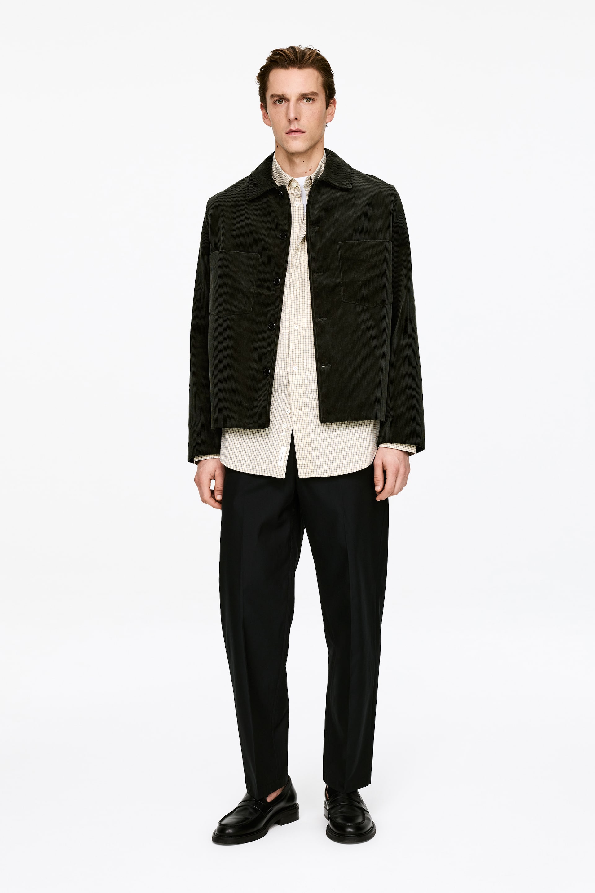 Größeres Bild ansehen: Eine dunkelgrüne Cordjacke wird von vorne präsentiert, mit einem spitzen Kragen, einer Knopfleiste und zwei großen aufgesetzten Taschen auf der Brust. Sie wird offen über einem hellen, mikromusterten Hemd getragen.