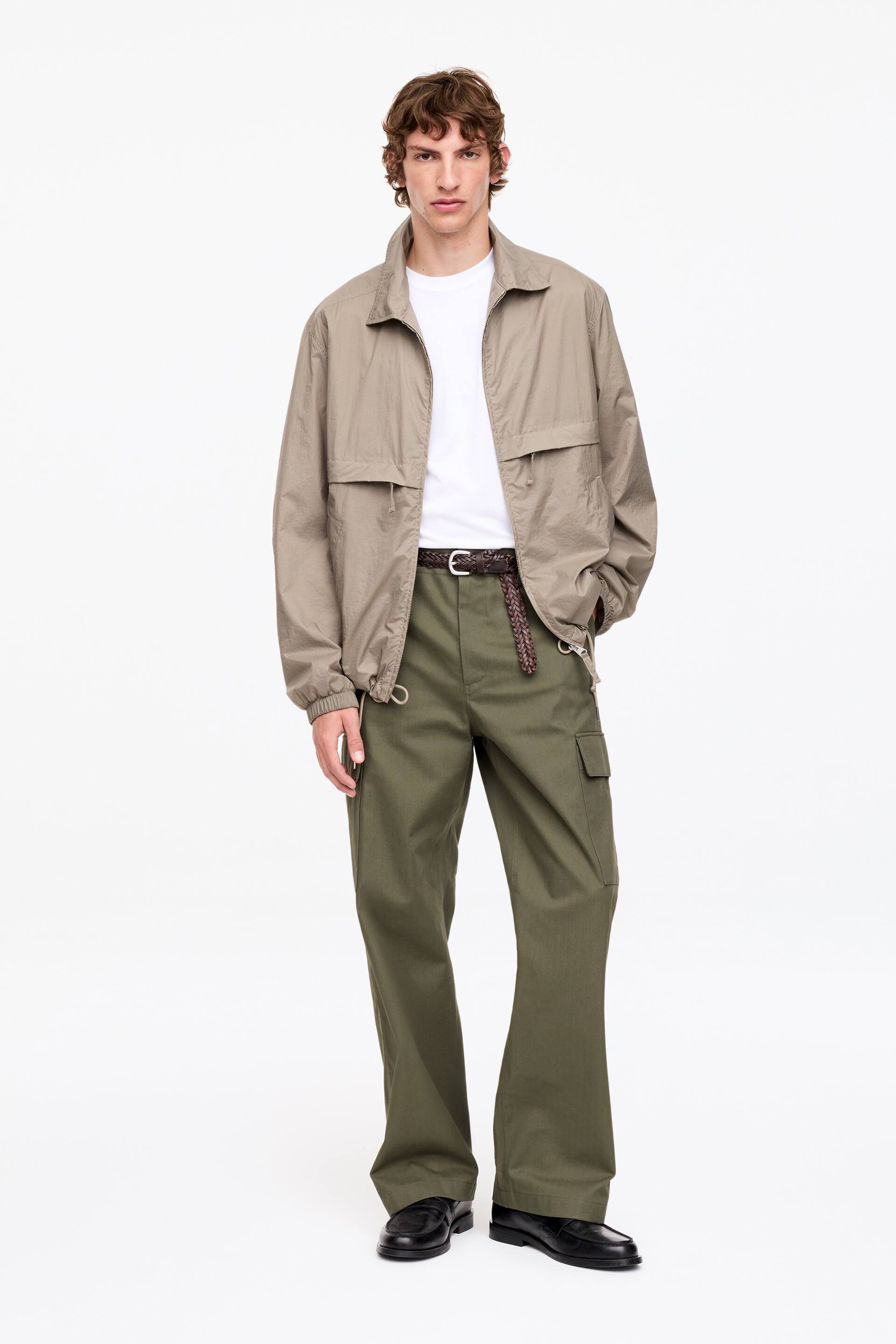 Agrandir l'image: Une veste légère taupe, dotée d'une fermeture éclair complète à l'avant, d'un col pointu et de deux poches à rabat horizontales, est portée ouverte sur un t-shirt à col rond blanc. Un pantalon cargo vert olive à jambes larges, avec des poches cargo latérales et maintenu par une ceinture en cuir tressé marron foncé, complète l'ensemble avec des mocassins en cuir noir, tous orientés vers l'avant.