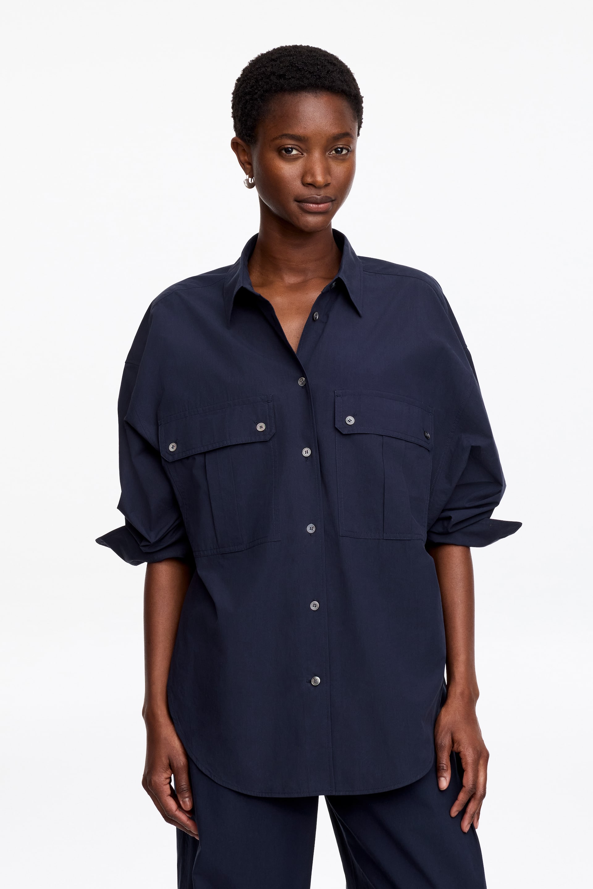 Agrandir l'image: Une chemise bleu marine foncé, arborant une silhouette ample, est portée par le mannequin. Elle présente une fermeture à boutons sur le devant, deux poches poitrine avec plis creux et fermetures à boutons, et ses manches longues sont retroussées.