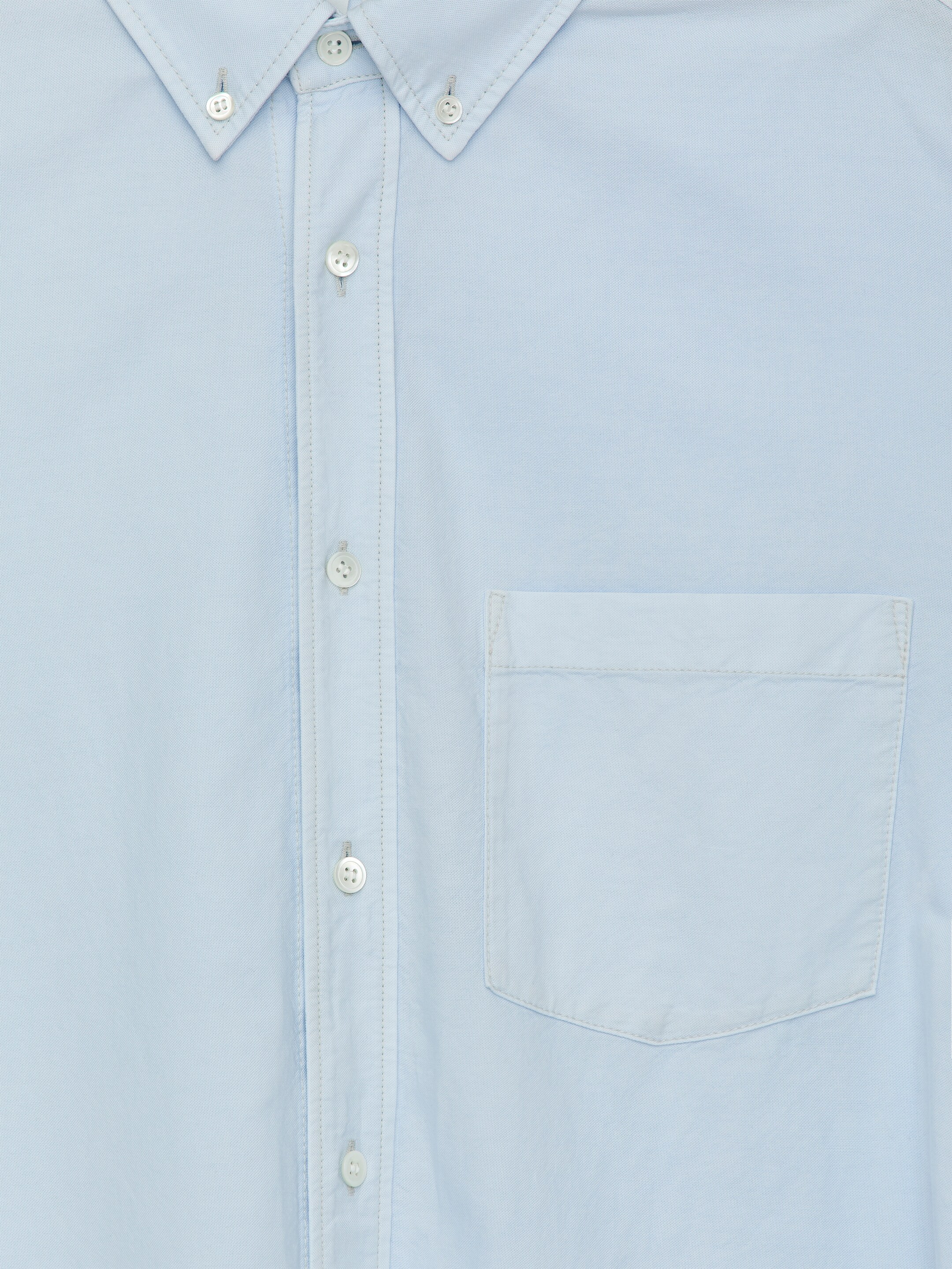 Agrandir l'image: Le DEVANT d'une chemise teintée en bleu clair, confectionnée dans un tissage coton Oxford, présente un col boutonné, une patte de boutonnage centrale avec des boutons en nacre et une poche poitrine à rabat unique.