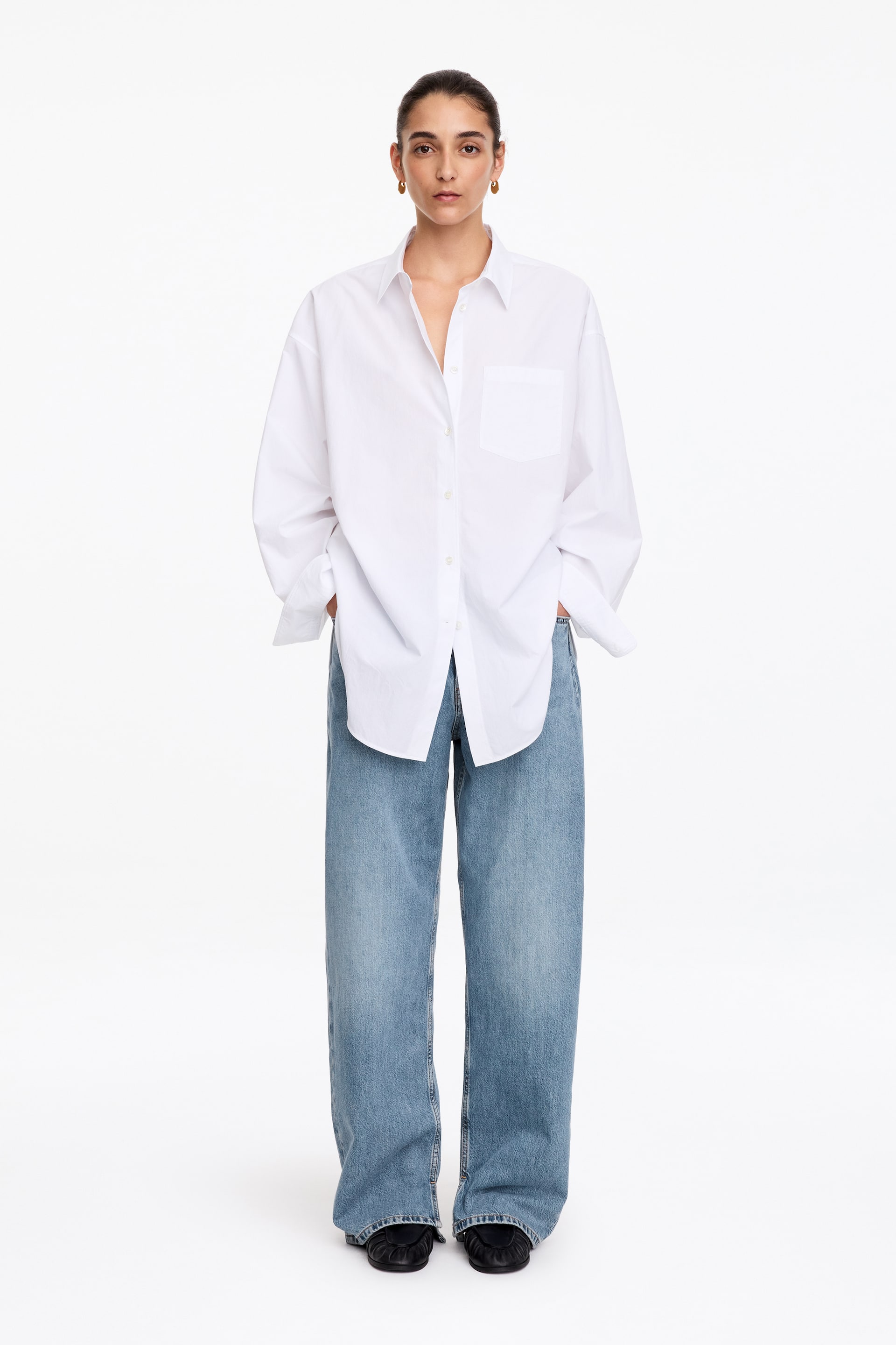Grotere afbeelding bekijken: De model draagt lichtblauwe jeans met een relaxed fit, gecombineerd met een oversized wit overhemd met knoopsluiting dat los over de broek valt. De jeans hebben een wide-leg silhouet en een klassieke denim wassing.
