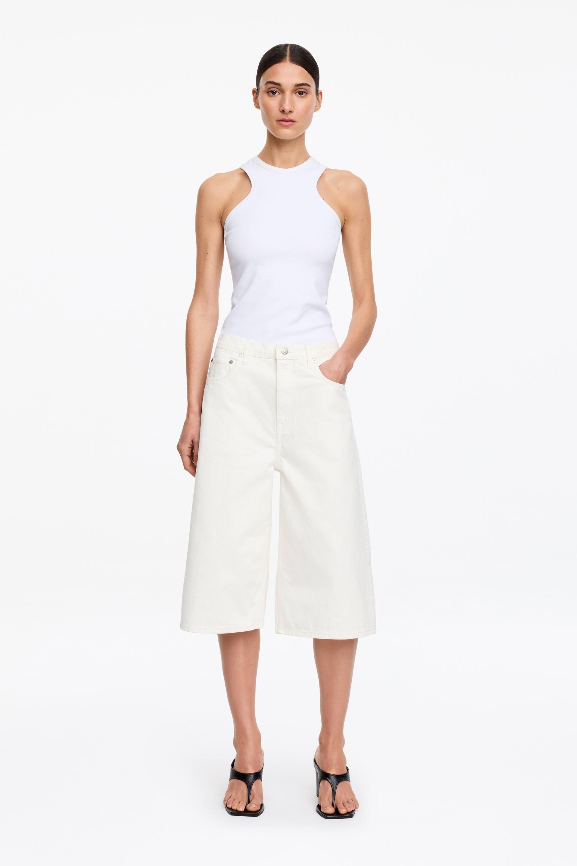 Ver imagen más grande: La modelo lleva unos pantalones cortos de denim color blanco roto, cortados a la altura de la rodilla con una silueta relajada. Presentan una construcción de cinco bolsillos, una bragueta con cremallera y un botón de remache. La modelo coloca su mano izquierda en un bolsillo delantero mientras los combina con una camiseta de tirantes de canalé blanca y sandalias de tacón alto negras.