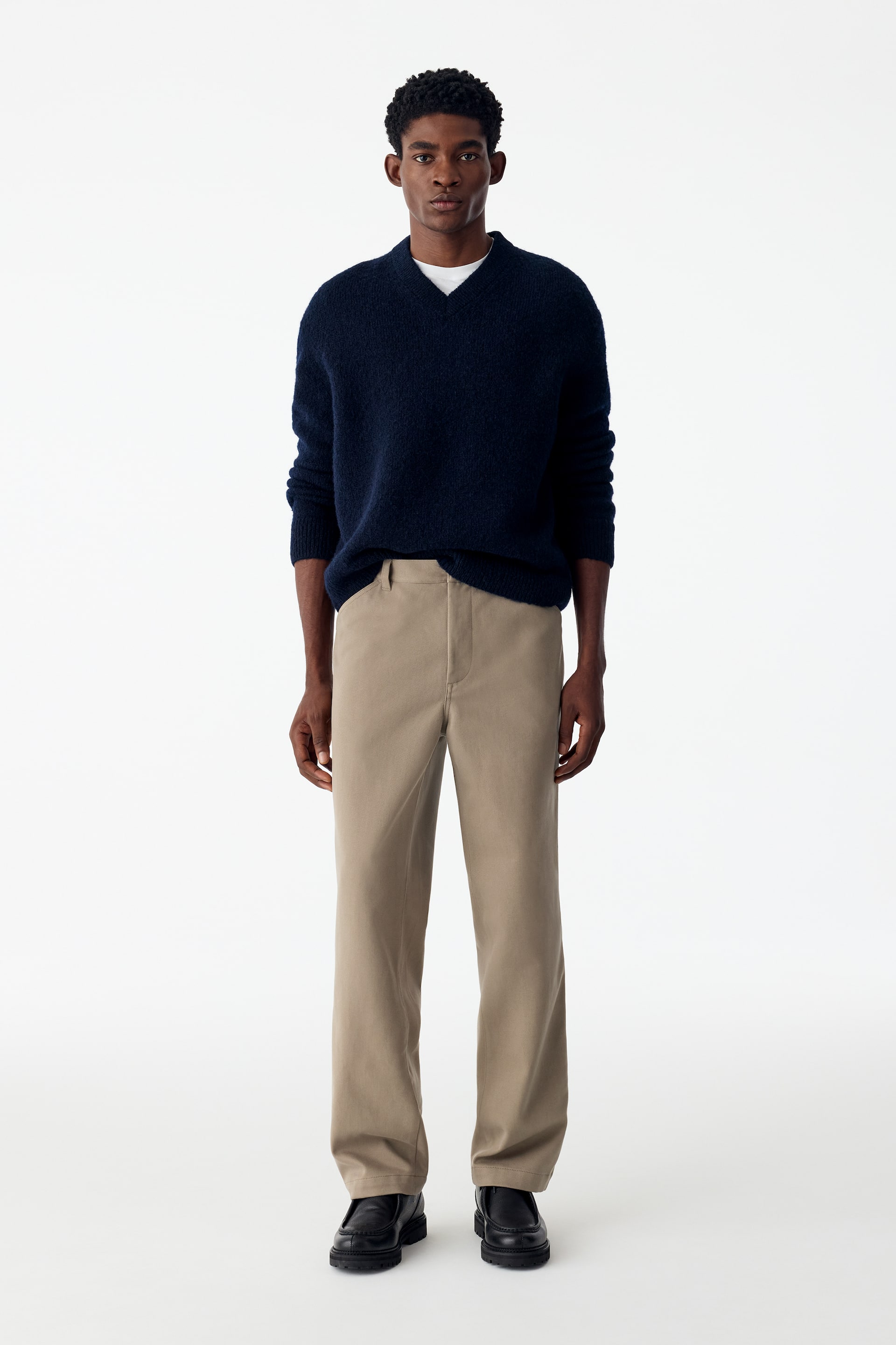 Grotere afbeelding bekijken: Relaxte katoenen lyocellbroek in beige wordt gedragen door het model, met een rechte pijp en een strakke tailleband. De broek is gestyled met een marineblauwe V-halstrui die gedeeltelijk in de voorzijde van de tailleband is ingestopt en gecombineerd met zwarte leren schoenen.