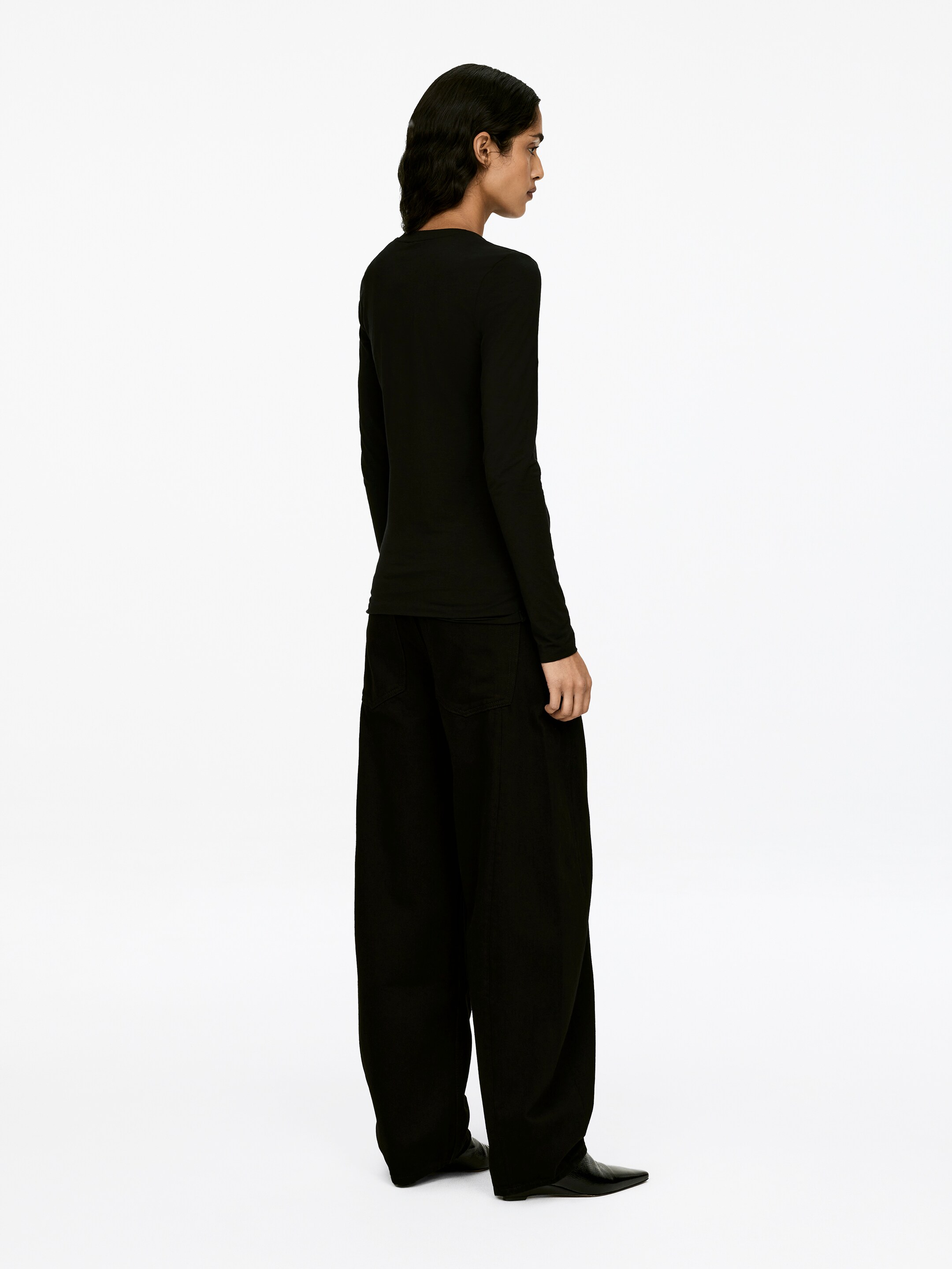 View larger image: A black long-sleeved top, described as soft and stretchy with a round-neck style and raw edges at the bottom hem and sleeve ends, is being worn by the model. The model is styling the top with black wide-leg pants and pointed-toe shoes.