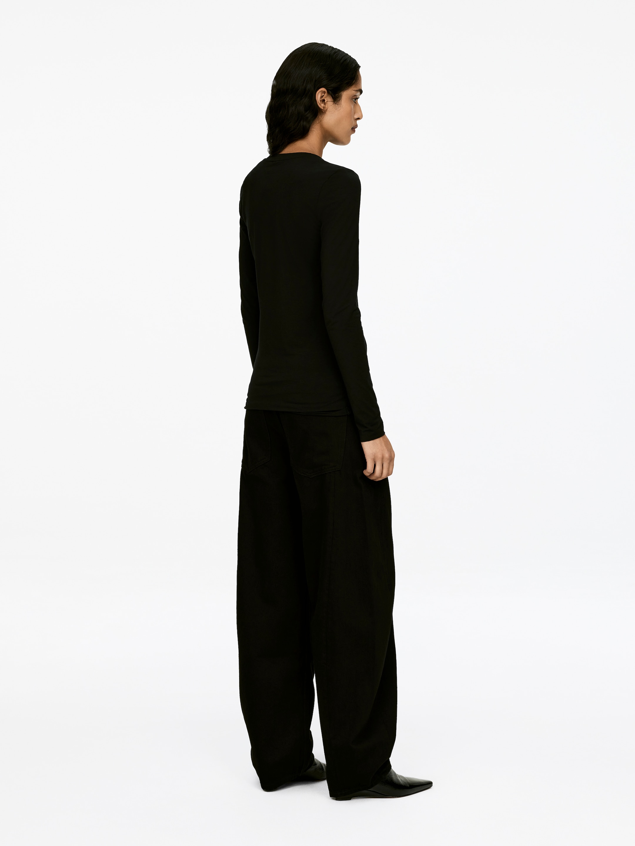 Se større billede: En sort langærmet top, beskrevet som blød og elastisk med en rund halsudskæring og rå kanter ved den nederste søm og ærmeenderne, bæres af modellen. Modellen styler toppen med sorte bukser med vidde ben og spidse sko.