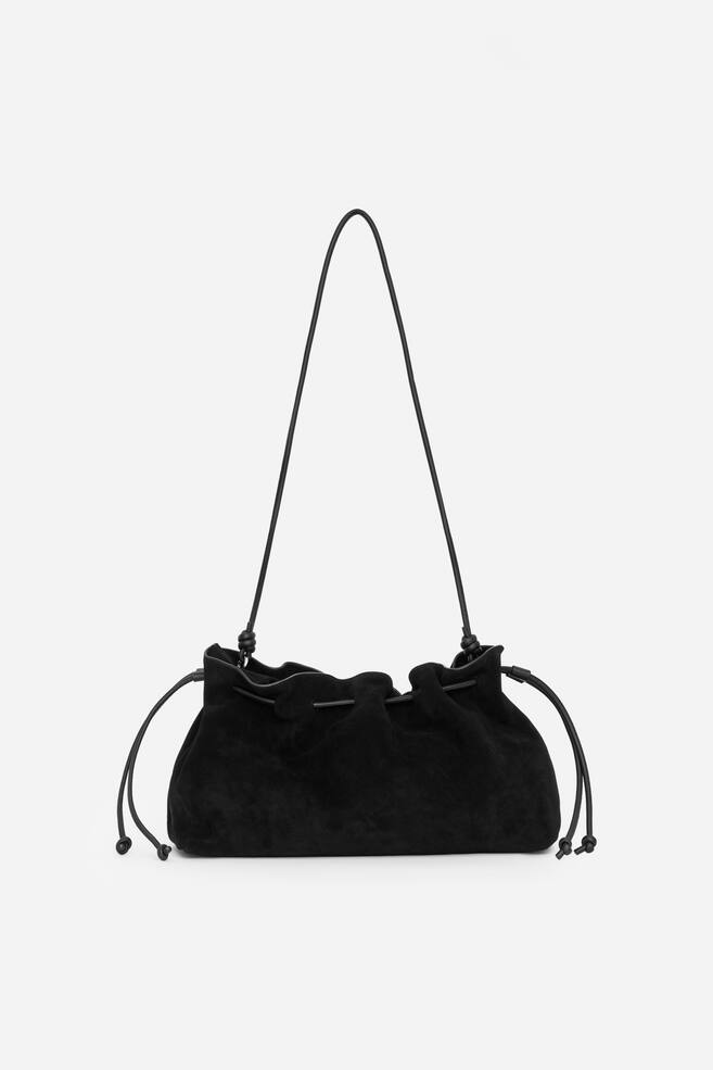 A black suede drawstring pouch bag is presenting its front, featuring a gathered top closure cinched by thin cords that extend to form a shoulder strap. - 1