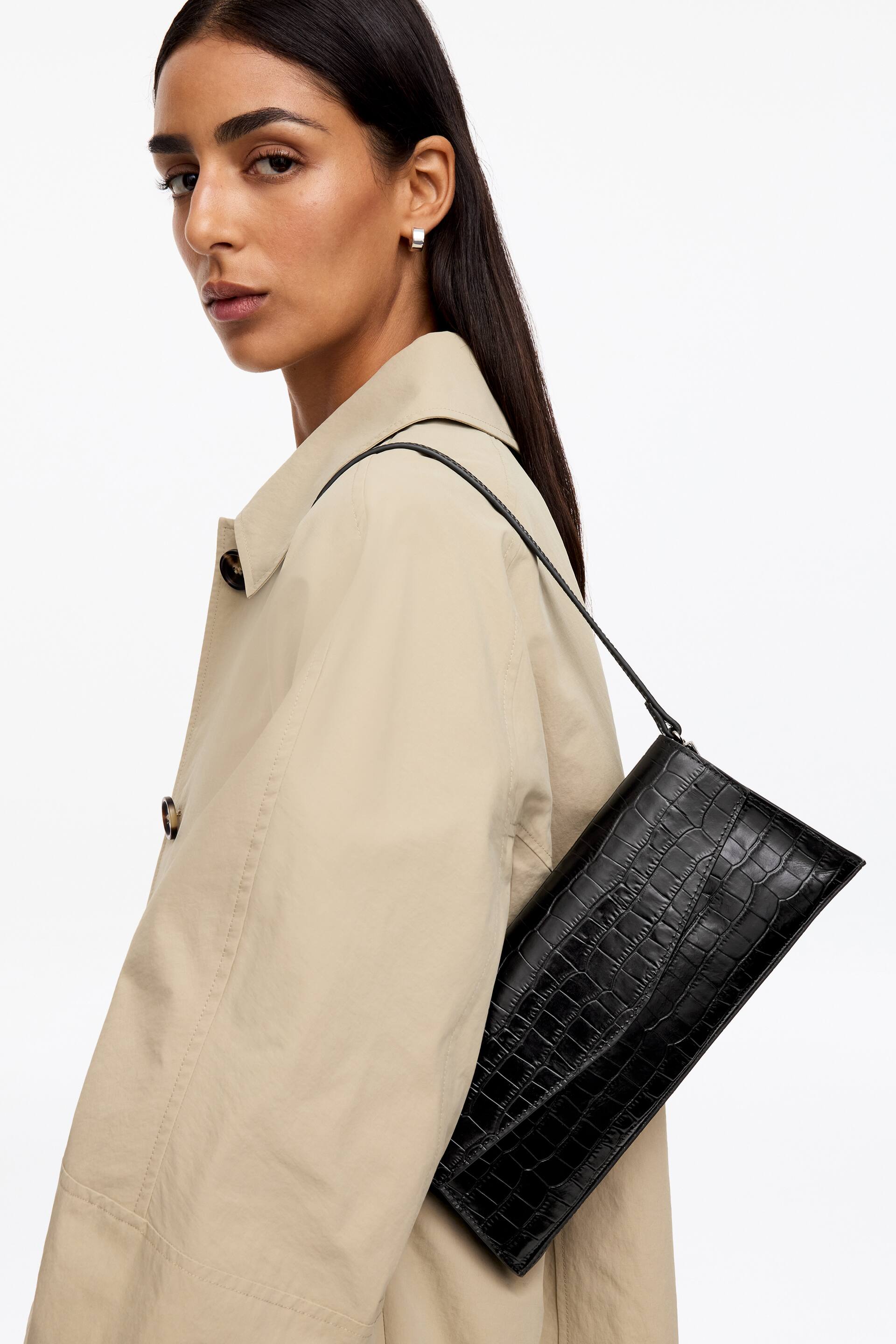 Näytä suurempi kuva: Nahkainen clutch-laukku. Mallilla on kädessään musta, suorakulmainen, krokotiilikuvioitu nahkainen clutch-laukku. Sen ohut hihna lepää hänen olkapäällään, ja hän pitää laukkua kylkeään vasten vaalean takin päällä.