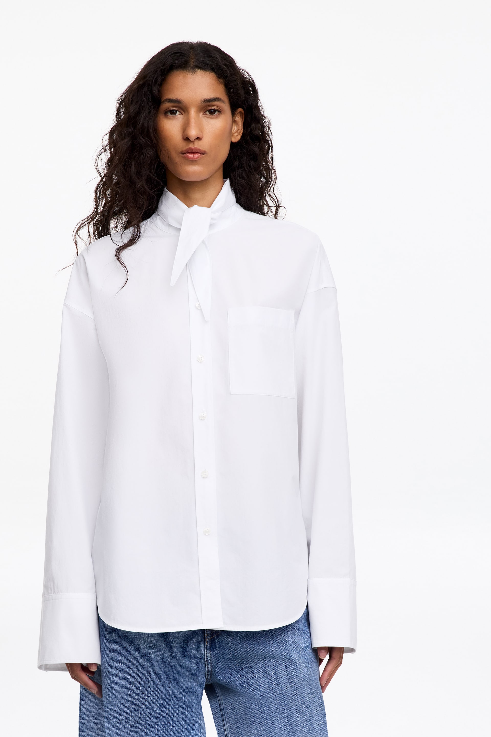 View larger image: The model is wearing a white long-sleeved shirt featuring a stand collar with front tie detail, a buttoned placket, a chest pocket, and wide cuffs. The shirt is styled untucked over blue jeans.