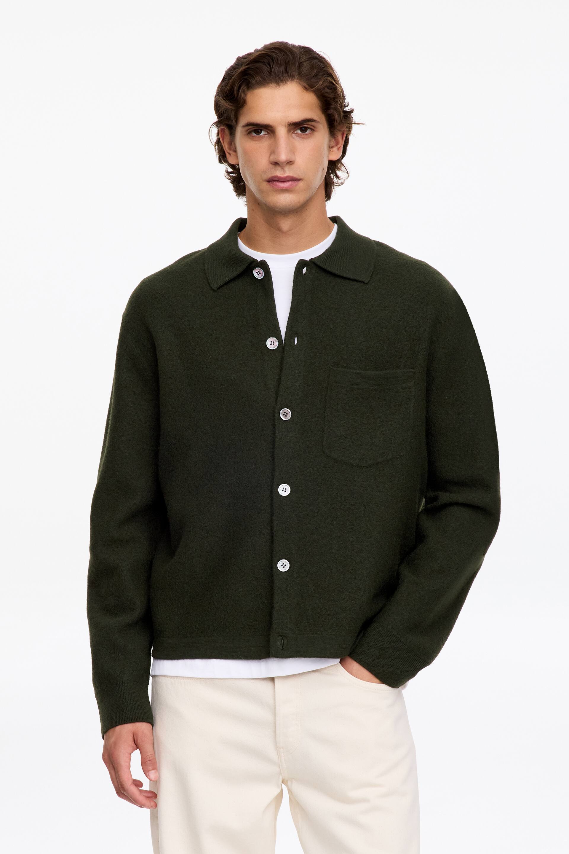View larger image: A dark green knitted collared jacket with a button-front fastening and a single chest patch pocket is being worn by the model. The jacket is layered over a white crew neck T-shirt and paired with off-white trousers.