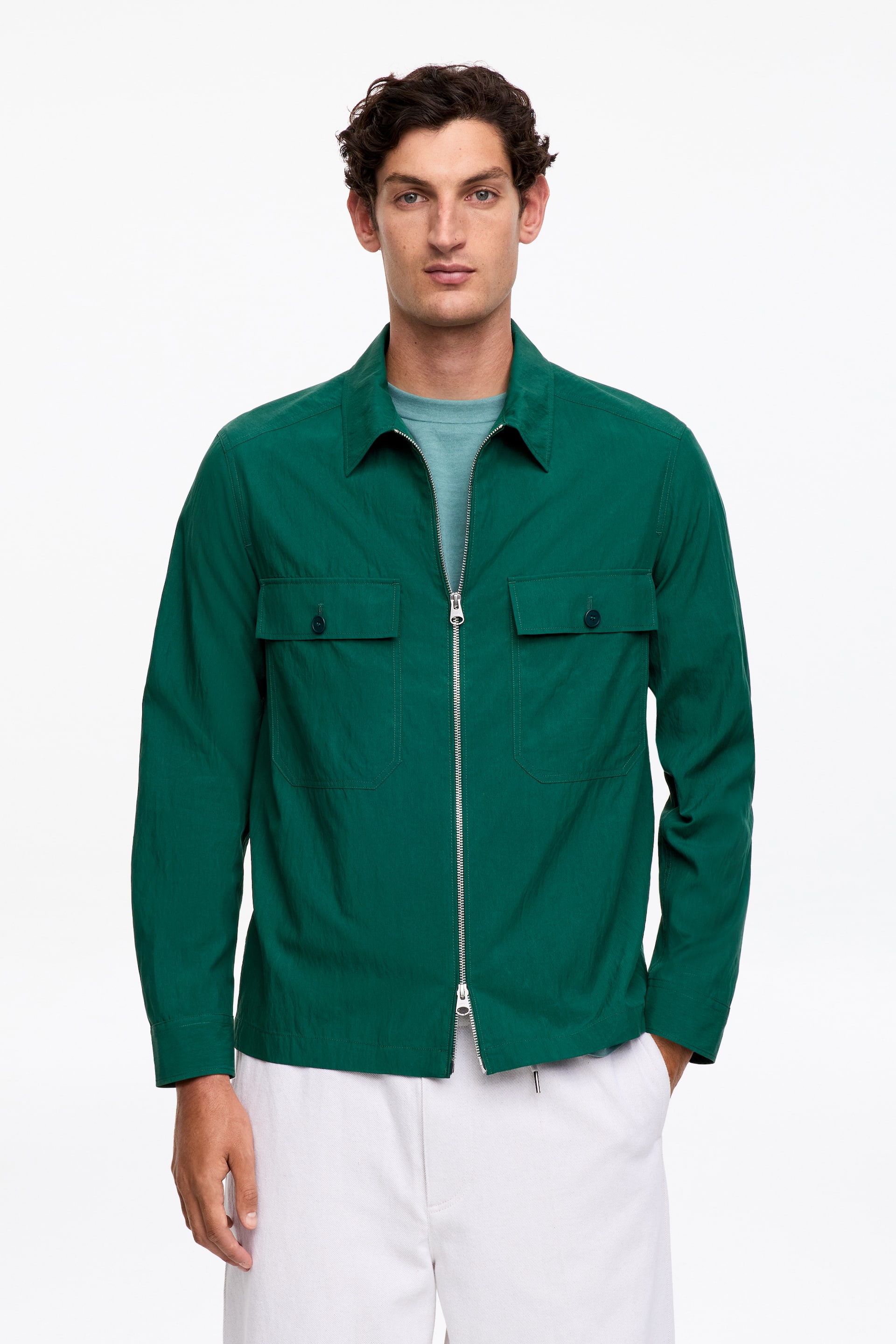 Zobrazit větší obrázek: Model má na sobě tmavě zelenou košili s pravidelným límcem, částečně rozepnutým dvoucestným předním zipem a náprsními kapsami s klopami na knoflíky, kterou nosí přes světle zelené tričko.