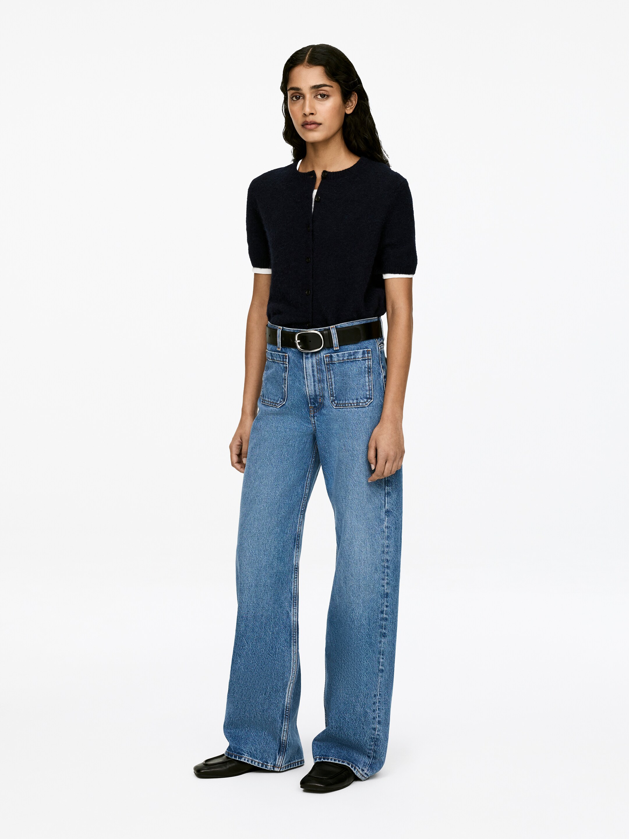 Agrandir l'image: Le modèle porte les jeans LUPINE. Une taille haute, une coupe ajustée au niveau des cuisses et des jambes évasées façonnent la silhouette. Des poches plaquées sur le devant sont visibles sur les jeans, et une ceinture noire serre la taille.