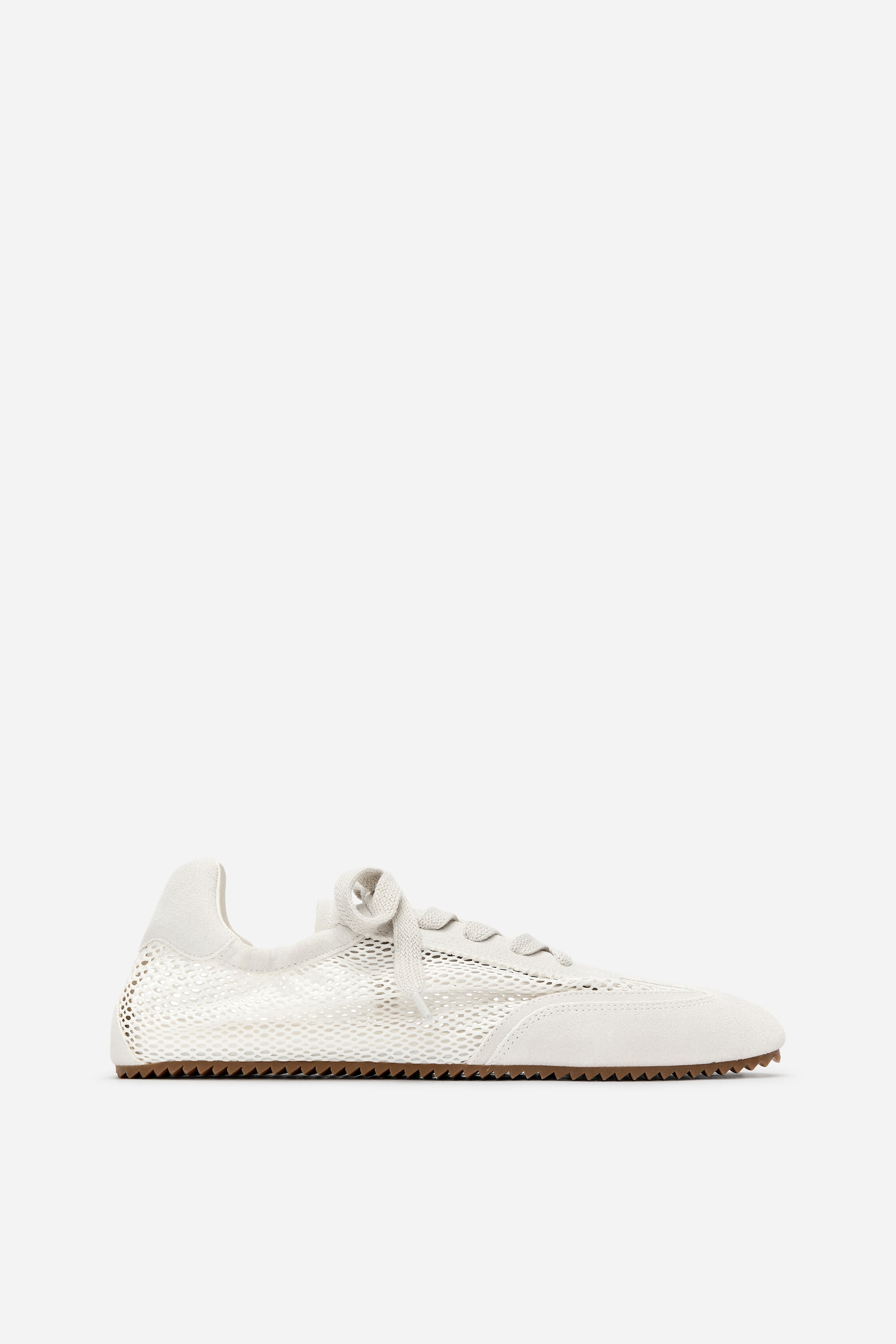 View larger image: An off-white trainer is shown from its left side, featuring a mesh upper with suede-like overlays on the toe, eyestays, and heel. Matching laces are tied in a bow, and a thin, serrated brown sole is visible beneath.