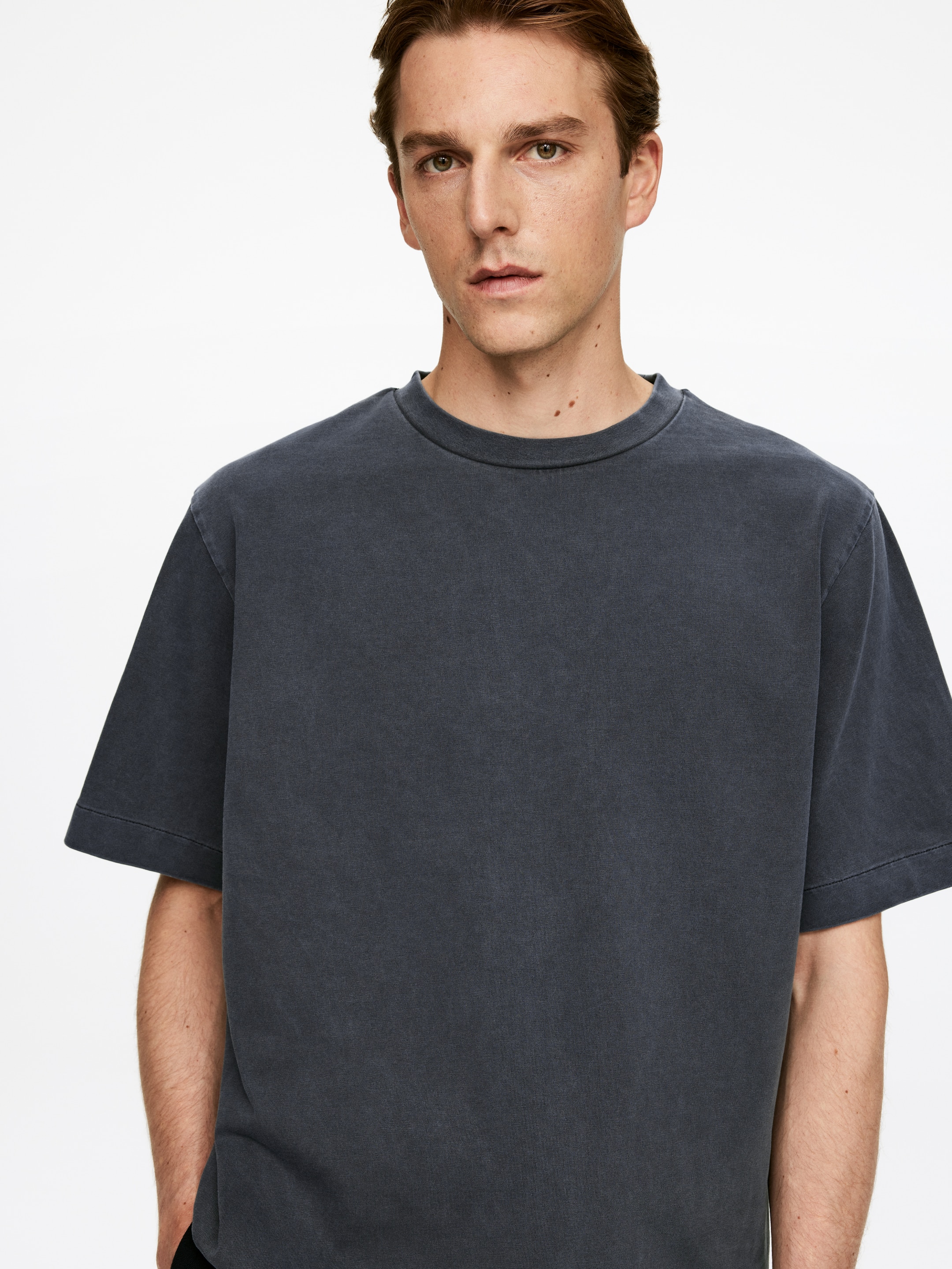 Ver imagen más grande: Una camiseta gris oscuro se presenta de frente, con un ajuste relajado. La camiseta está diseñada con un cuello redondo acanalado y mangas cortas y anchas, mostrando una textura visible lavada o descolorida.
