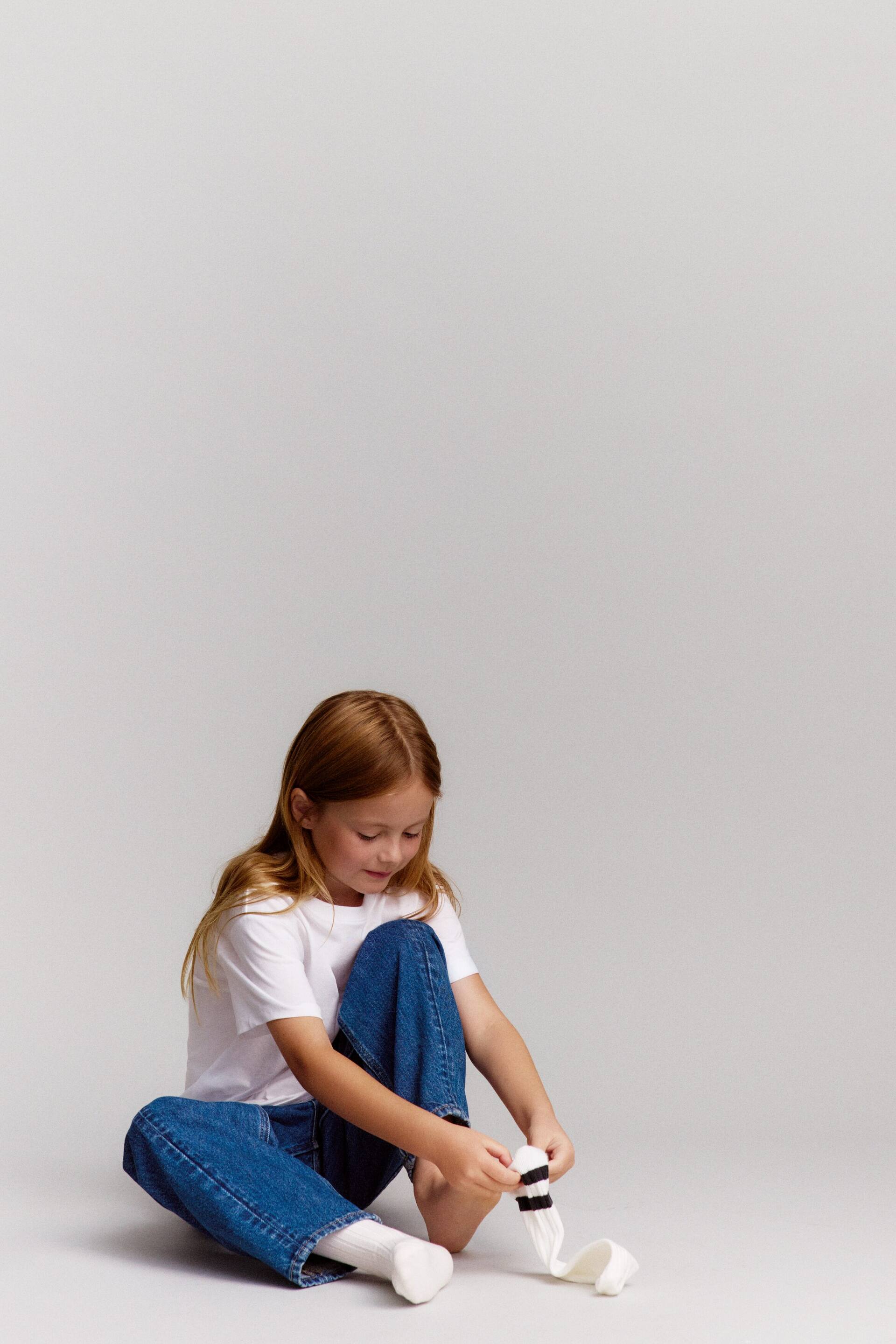 Grotere afbeelding bekijken: Een kind is van voren te zien, gekleed in een witte T-shirt met ronde hals en regular fit, en een blauwe barrel jeans met een wijde, sculpturale silhouet. Het zit terwijl het een witte geribbelde sok met zwarte strepen op de schacht aantrekt.