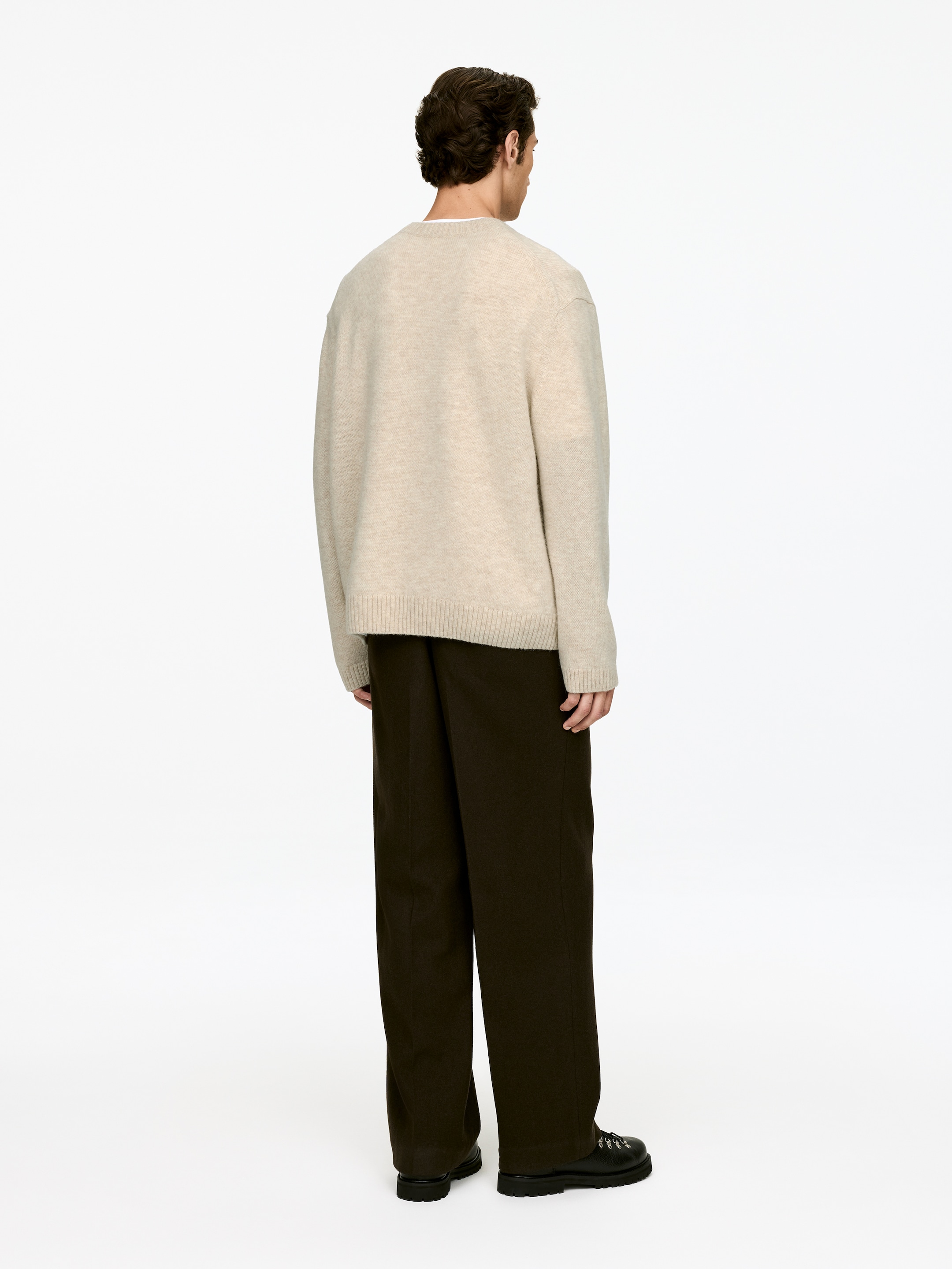 Visa större bild: En beige tröja med rund hals visas bakifrån och visar dess avslappnade passform och ribbade fåll. Den bärs med mörkbruna, vida byxor som faller över svarta kraftiga snörstövlar.