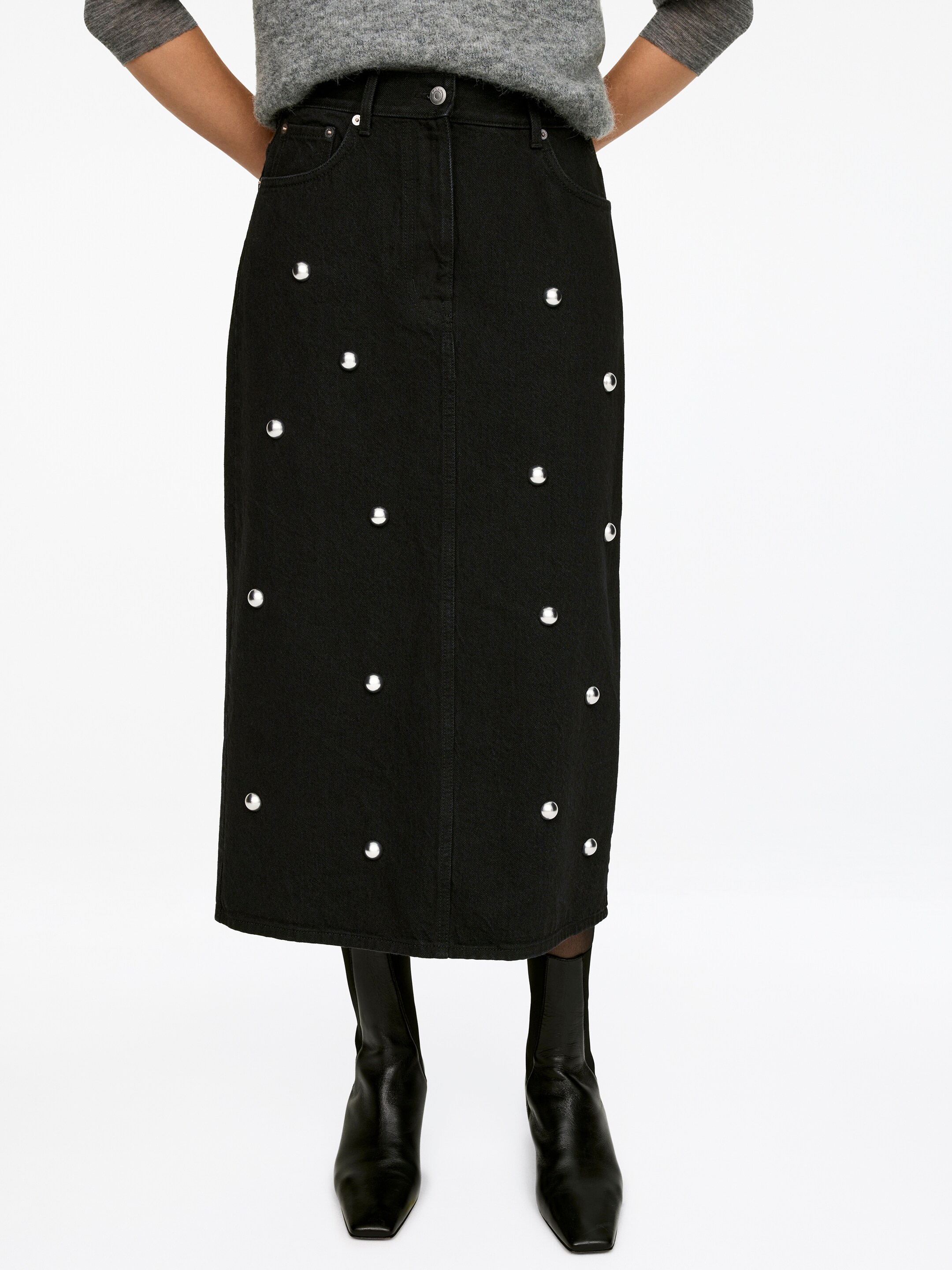 Agrandir l'image: Une jupe noire en coton de longueur moyenne, avec un design à cinq poches et ornée de clous argentés, est portée par le mannequin. La jupe a une coupe régulière et est assortie avec un pull gris et des bottes noires.