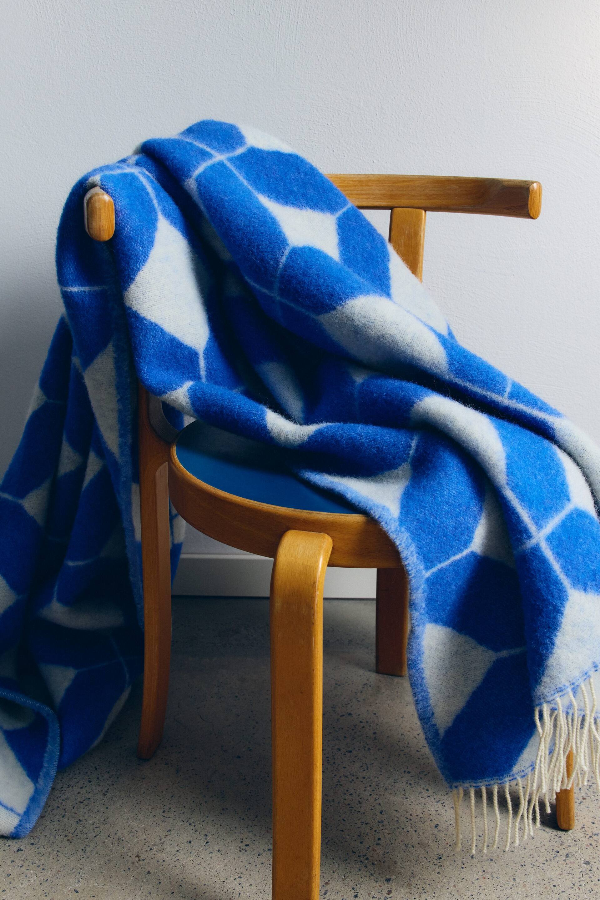 Agrandir l'image: Une couverture à losanges bleus et blancs est drapée sur une chaise en bois, avec des franges blanches clairement visibles le long d’un bord pendant.