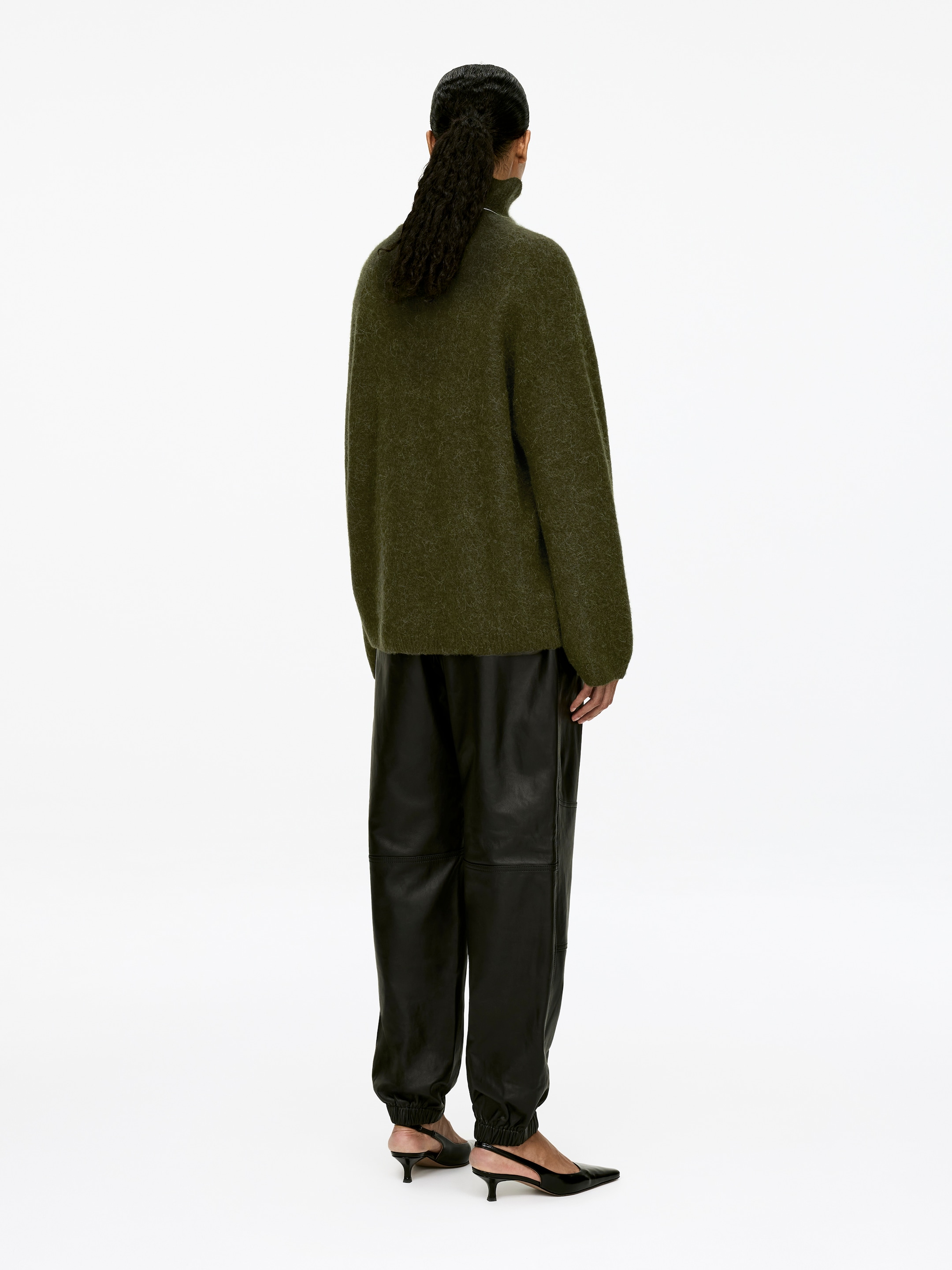 Agrandir l'image: Un pull en tricot duveteux à col montant vert olive avec des manches longues est porté, vu de dos, sur un pantalon jogging en cuir noir avec des poignets élastiques et des coutures visibles aux genoux.