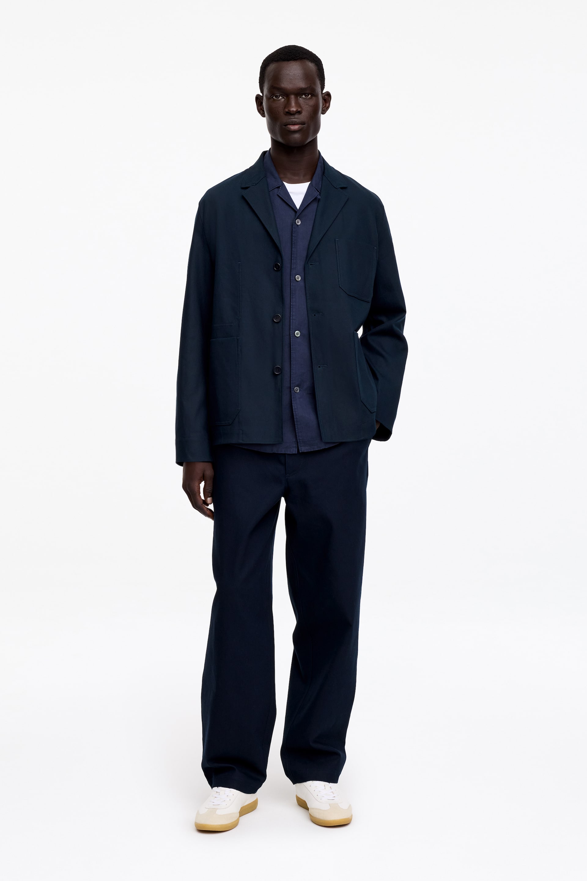 Zobrazit větší obrázek: Tmavě modrá bunda ve stylu pracovního oděvu je vybavena klasickým límcem, zapínáním na tři knoflíky vpředu a dvěma velkými našitými kapsami, vrstvená přes tmavě modrou košili s reverzním límcem a bílé tričko s kulatým výstřihem. Odpovídající tmavě modré kalhoty s širokými nohavicemi doplňují bundu spolu s bílými teniskami s béžovými semišovými akcenty a gumovými podrážkami.