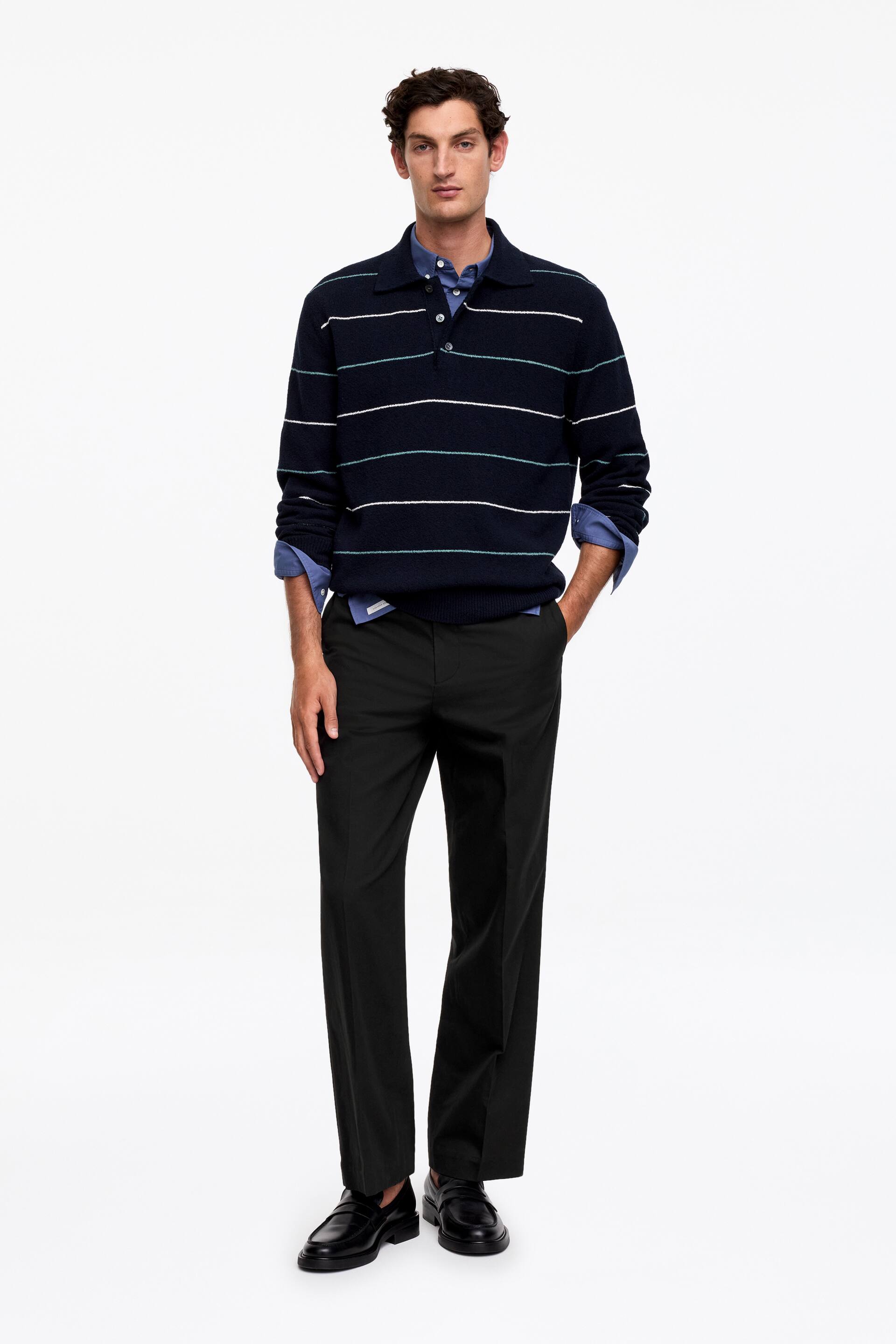 Grotere afbeelding bekijken: Een marineblauwe polotrui met horizontale witte en lichtblauwe strepen wordt gepresenteerd, gelaagd over een lichtere blauwe overhemd met kraag. De trui heeft een polokraag, een knoopsluiting met drie knopen, en geribbelde boorden en zoom, waarbij de kraag en manchetten van het overhemd zichtbaar uitsteken.