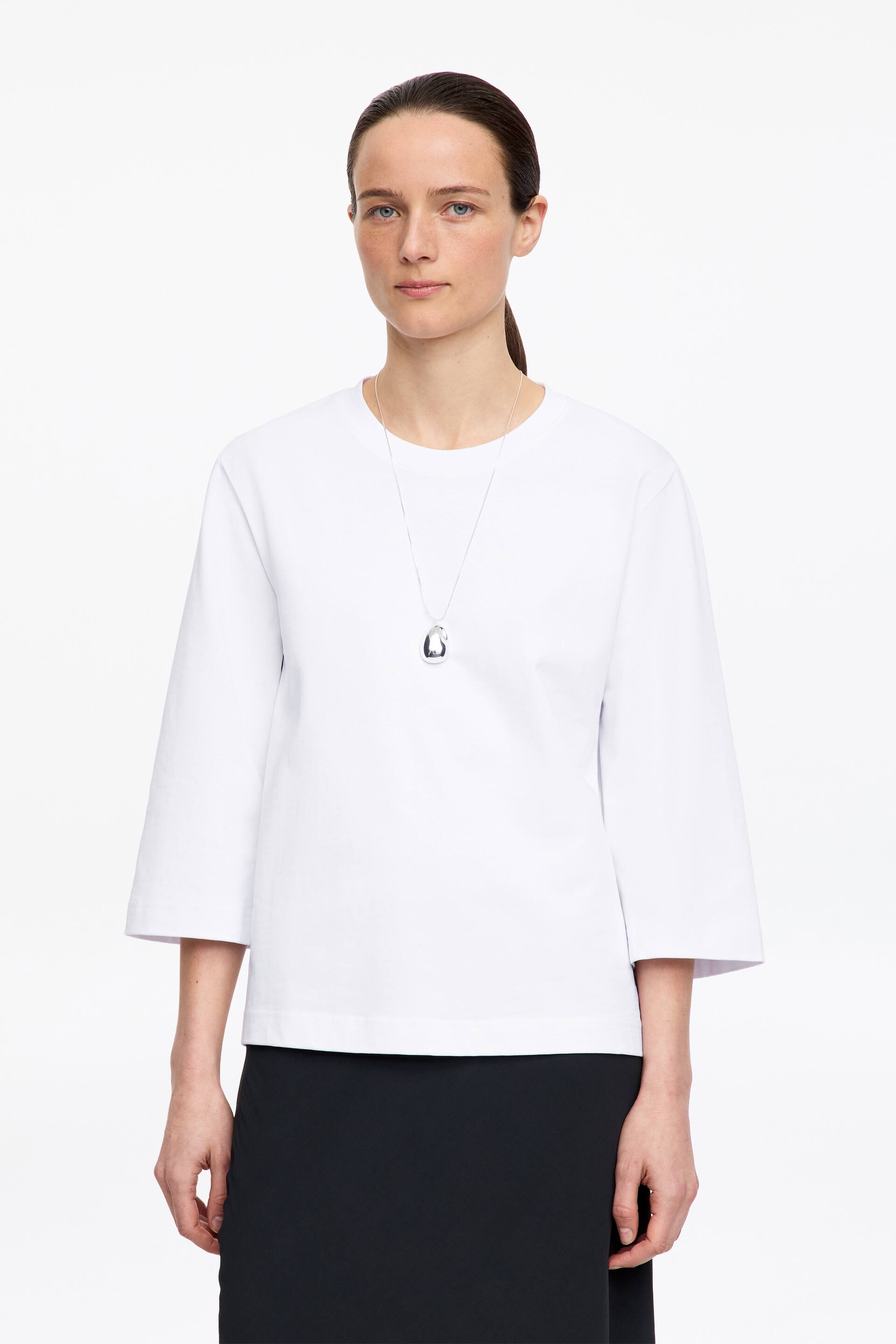 Größeres Bild anzeigen: Ein weißes strukturiertes Oberteil wird von vorne präsentiert, mit einem Rundhalsausschnitt und dreiviertellangen Ärmeln mit geraden Manschetten. Es fällt in einer entspannten Passform mit einem geraden Saum und wird über einem schwarzen Rock getragen, während eine silberne Anhängerkette über dem Oberteil getragen wird.