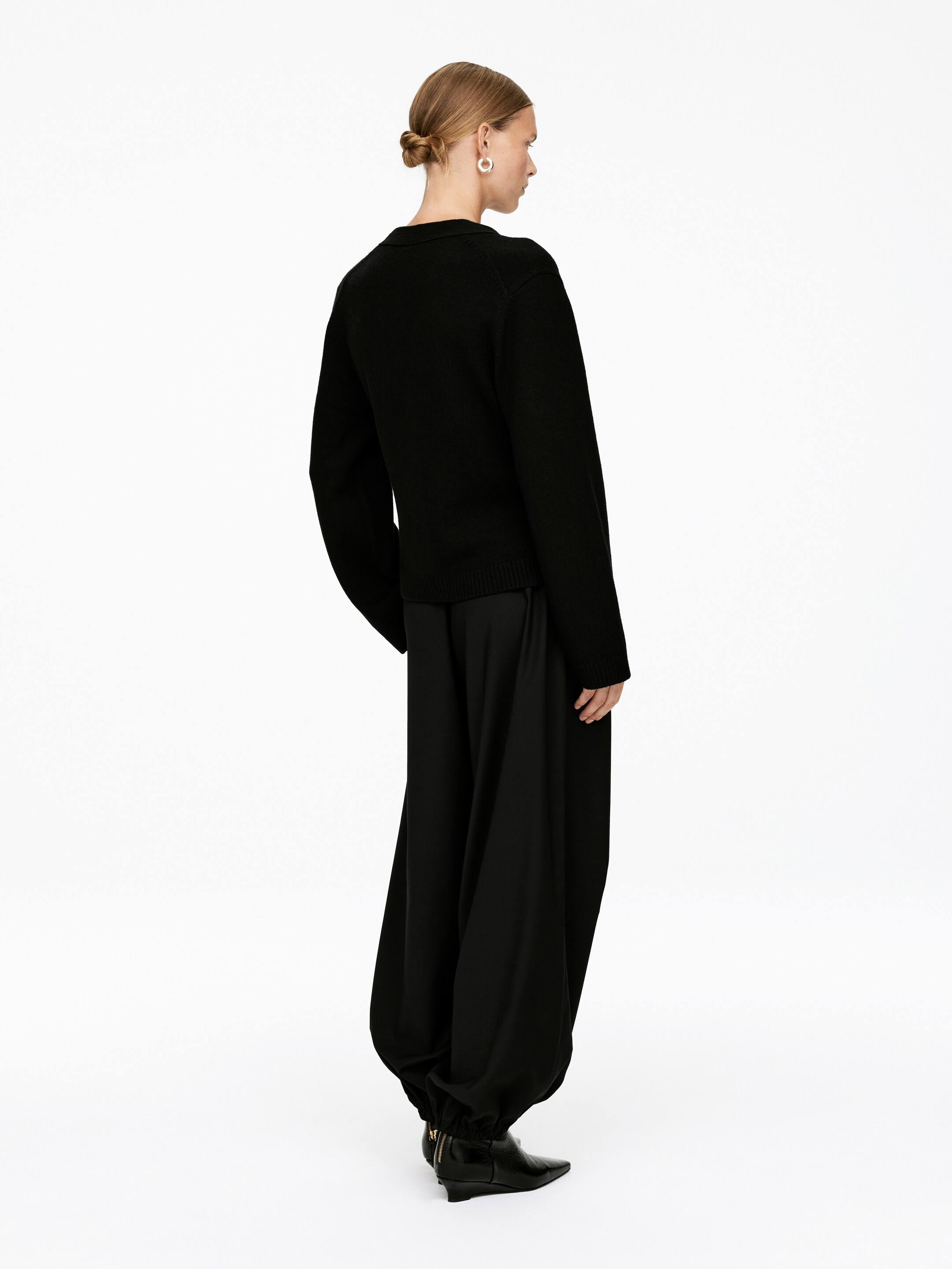 View larger image: A black knit sweater and black voluminous pants are being worn, viewed from a back and side angle. The sweater is featuring long sleeves and a ribbed hem, while the pants are creating a voluminous silhouette with wide legs that are cinching at the ankles, visibly styled over black pointed-toe ankle boots that are revealing a gold zipper.