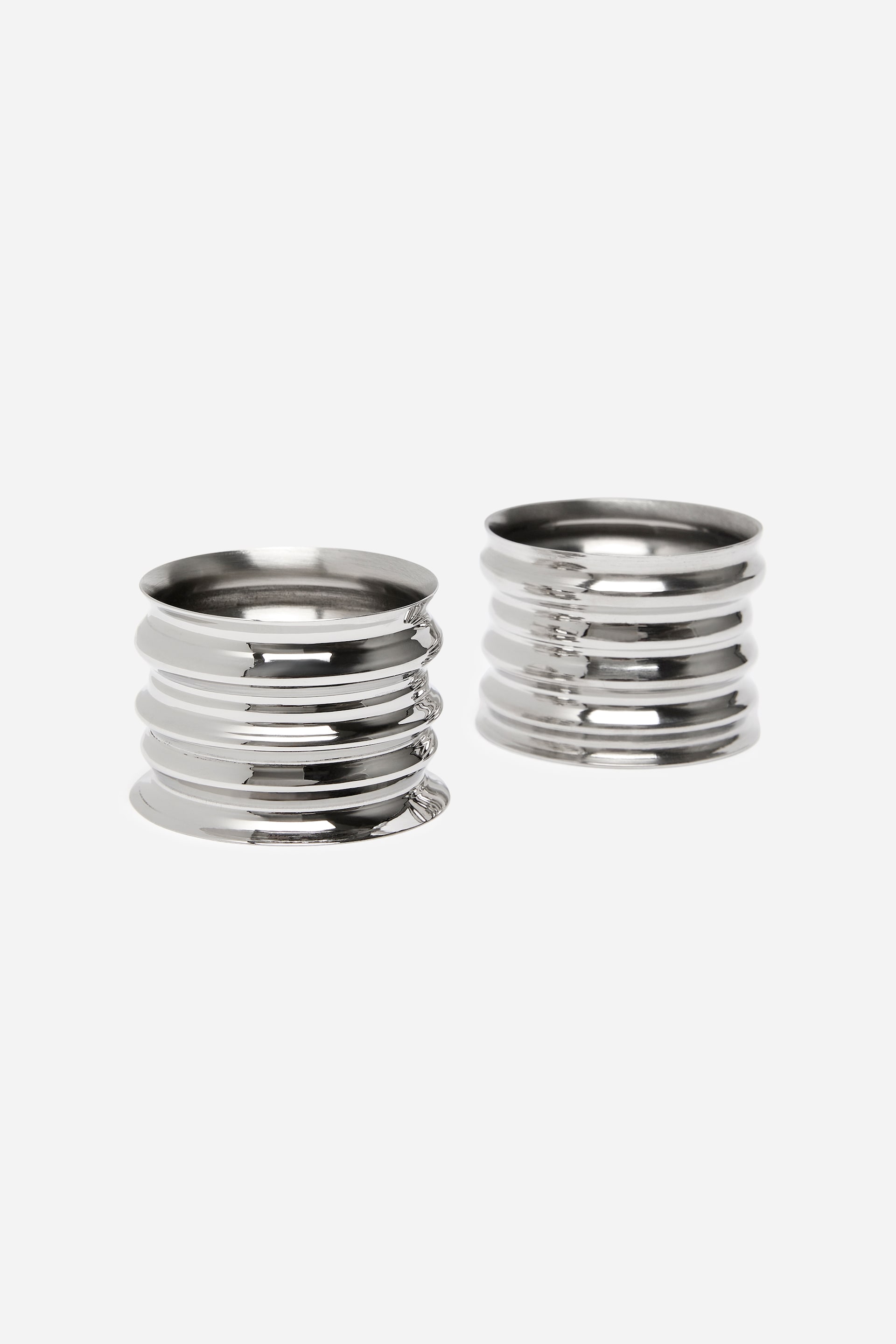 Vis større bilde: To sølvfargede metallserviettringer står side om side, med en polert, reflekterende overflate og et rillet, sylindrisk design med flere horisontale spor. Ringene er vist forfra, noe som fremhever de hule innsidene og de teksturerte ytterflatene.