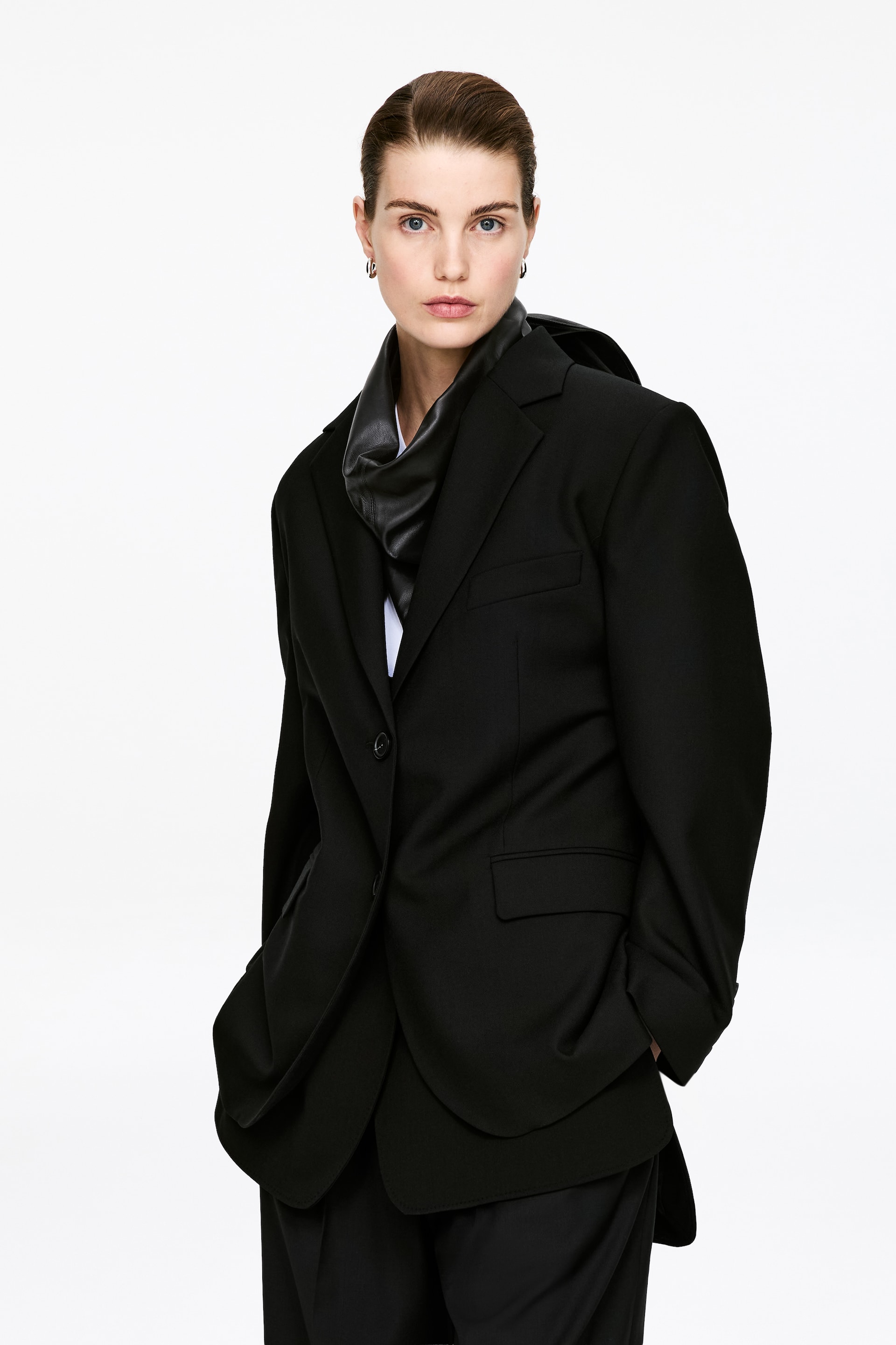 Zobrazit větší obrázek: Černé sako je zobrazeno z mírného úhlu, směřuje dopředu a doleva, vyznačuje se zářezovými klopami, zapínáním na jeden knoflík a klopovou kapsou na pravé straně. Černý doplněk kolem krku je vrstven pod jeho klopami.