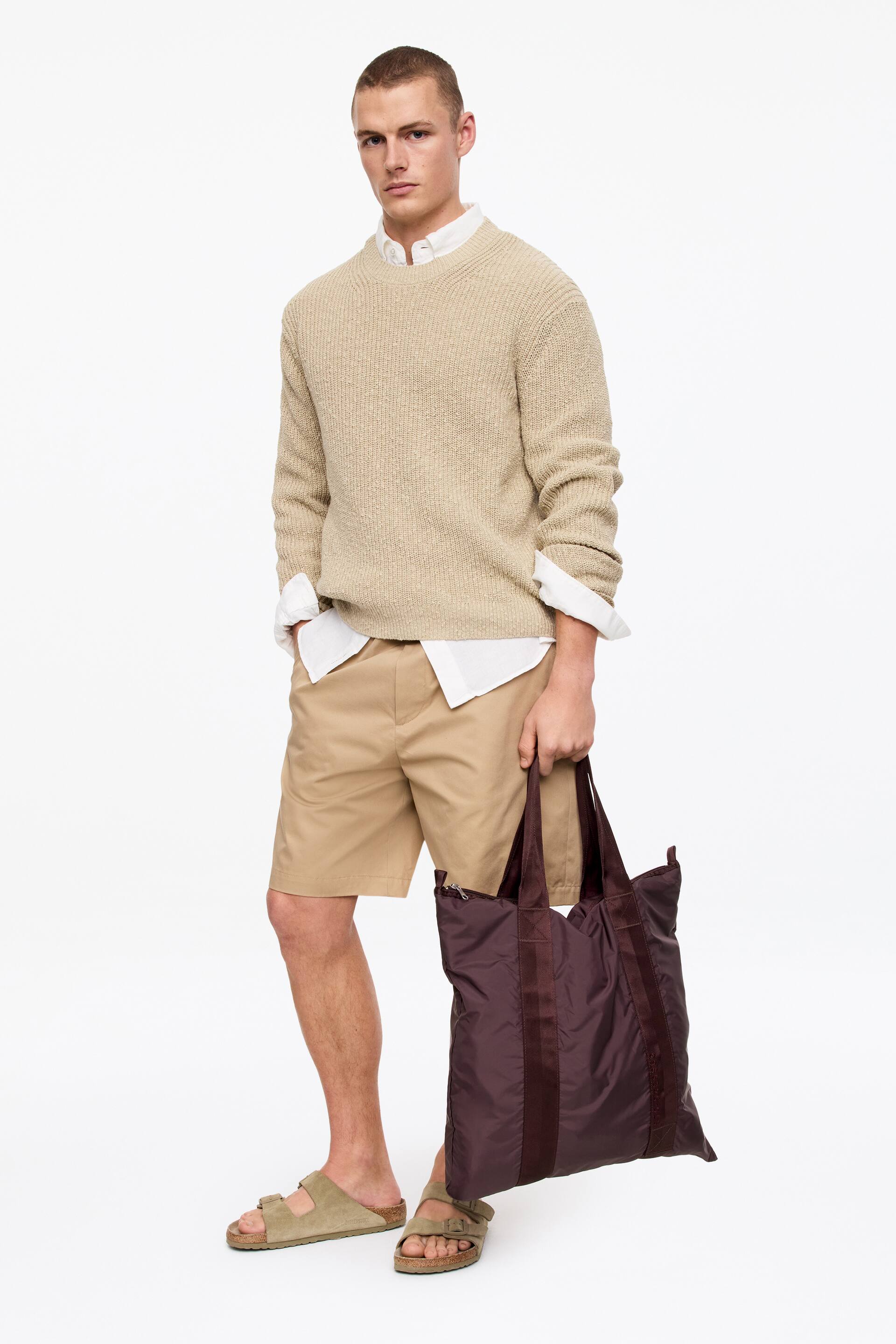 Näytä suurempi kuva: Tumma ruskea pakattava olkalaukku, valmistettu kevyestä, pehmeästä nailonkankaasta, jossa on kaksi litteää yläkahvaa ja pystysuuntainen koristehihna. Malli pitää olkalaukkua kahvoista, antaen sen roikkua sivullaan.