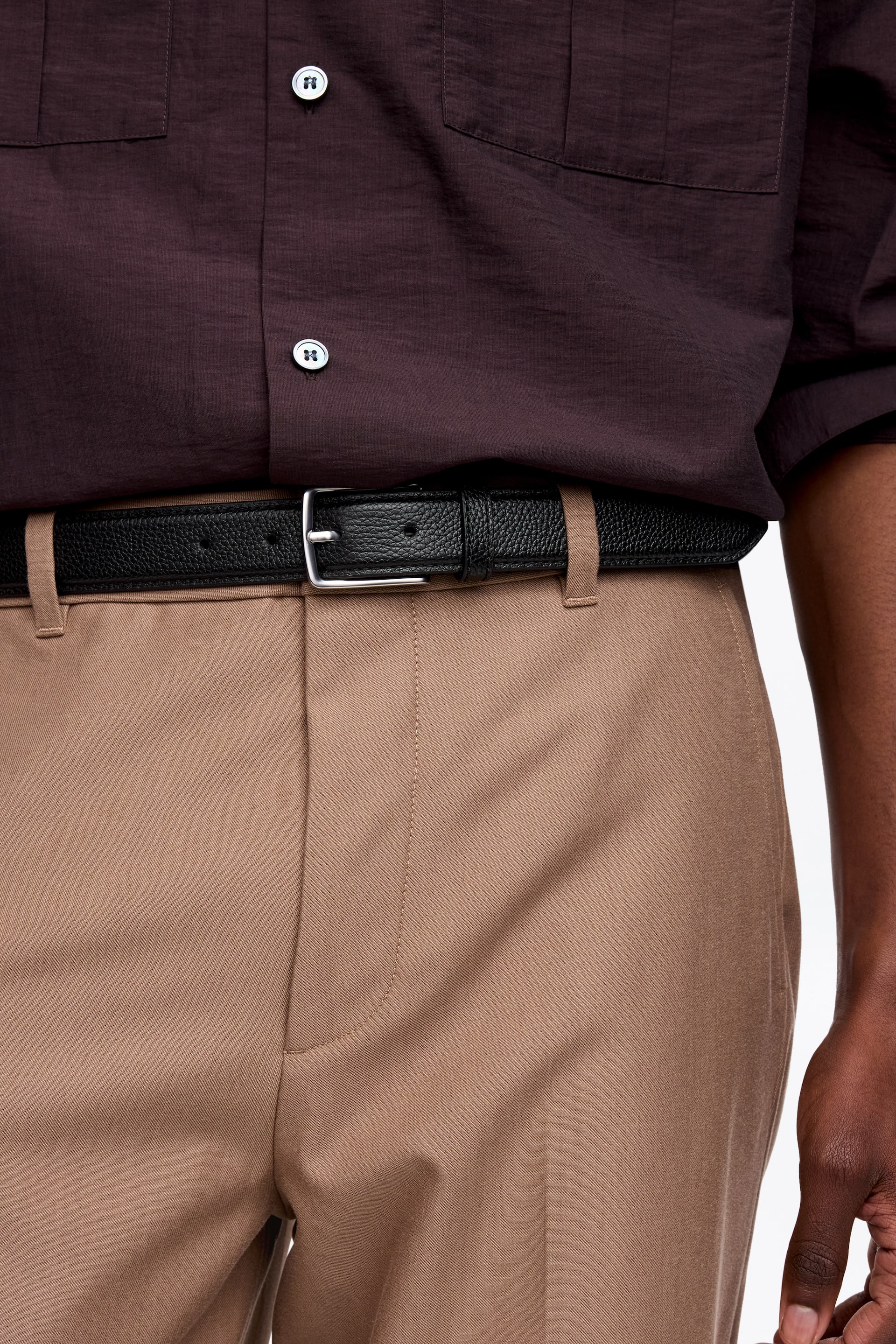 Προβολή μεγαλύτερης εικόνας: Μια μαύρη ζώνη από δέρμα με υφή βότσαλου σφίγγει ένα σκούρο καφέ πουκάμισο, φορεμένο μέσα από τις θηλιές ζώνης ενός ανοιχτού καφέ παντελονιού. Διαθέτει μια ορθογώνια αγκράφα σε ασημί τόνο.