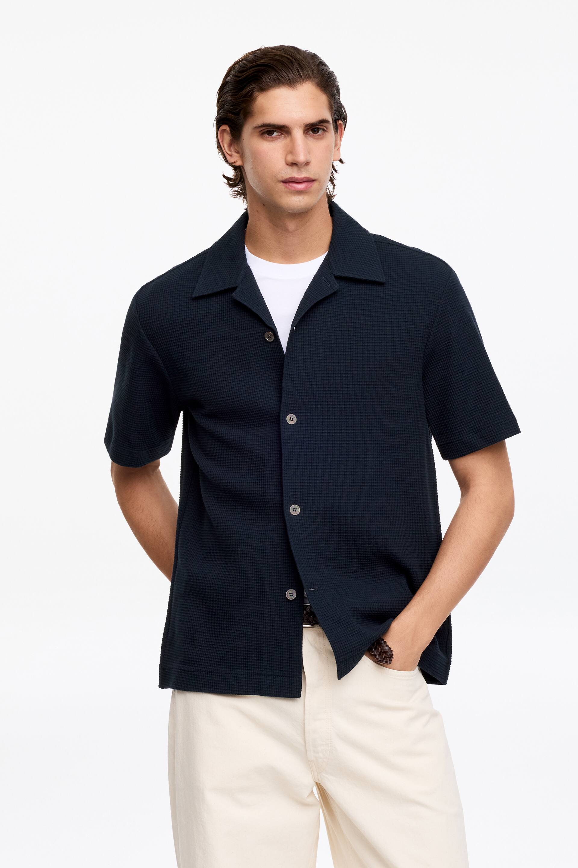 Προβολή μεγαλύτερης εικόνας: Το σκούρο μπλε πουκάμισο με πλέξη βάφλας και κοντά μανίκια διαθέτει γιακά τύπου camp και τρία ανοιχτόχρωμα κουμπιά κατά μήκος του μπροστινού μέρους, και παρουσιάζεται ανοιχτό πάνω από ένα λευκό t-shirt.