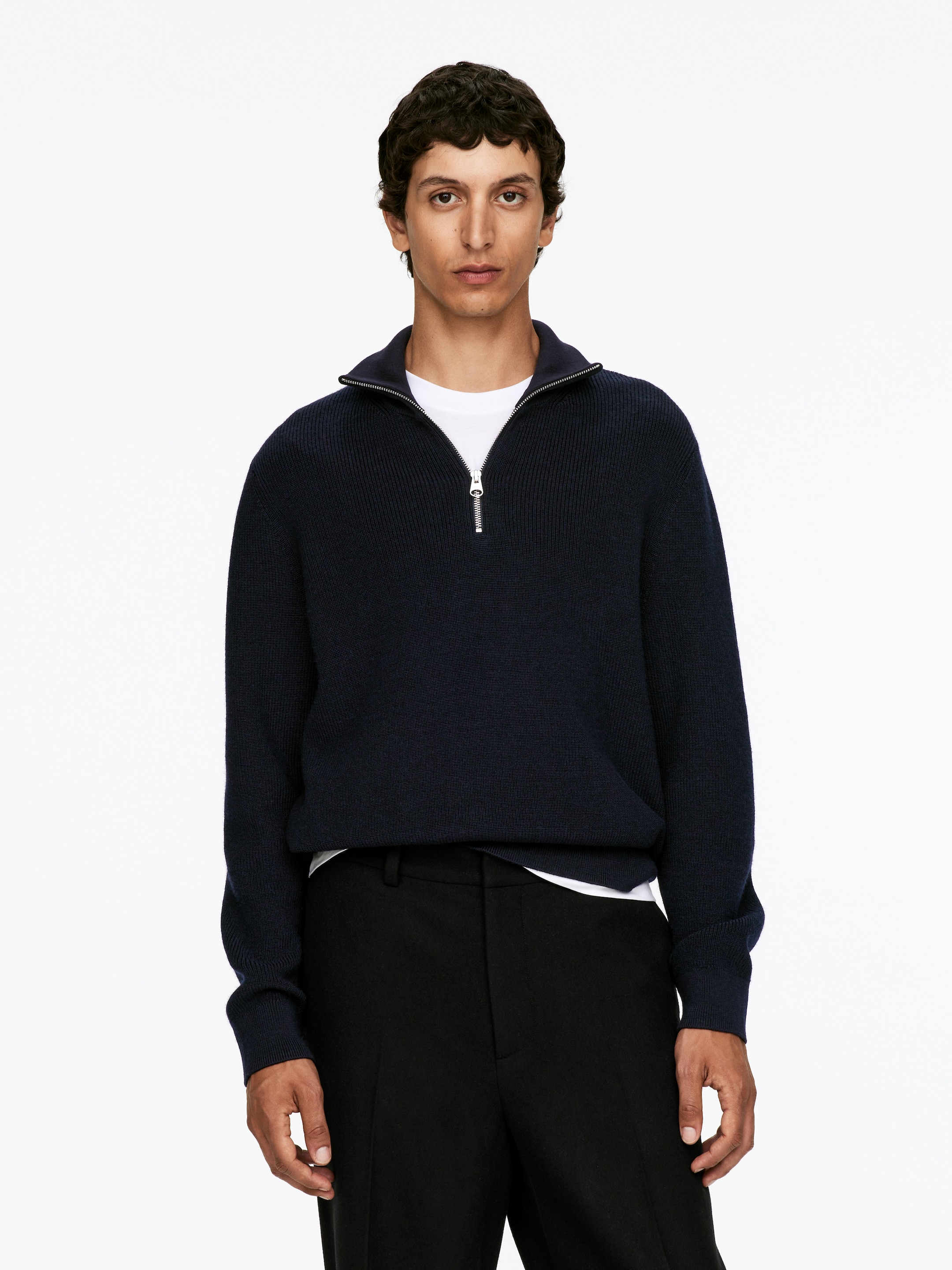 Visualizza immagine più grande: Un maglione a mezza zip blu scuro è indossato dal modello, caratterizzato da un colletto a coste e una cerniera argentata che è parzialmente aperta. Una t-shirt bianca con scollo a girocollo è stratificata sotto.