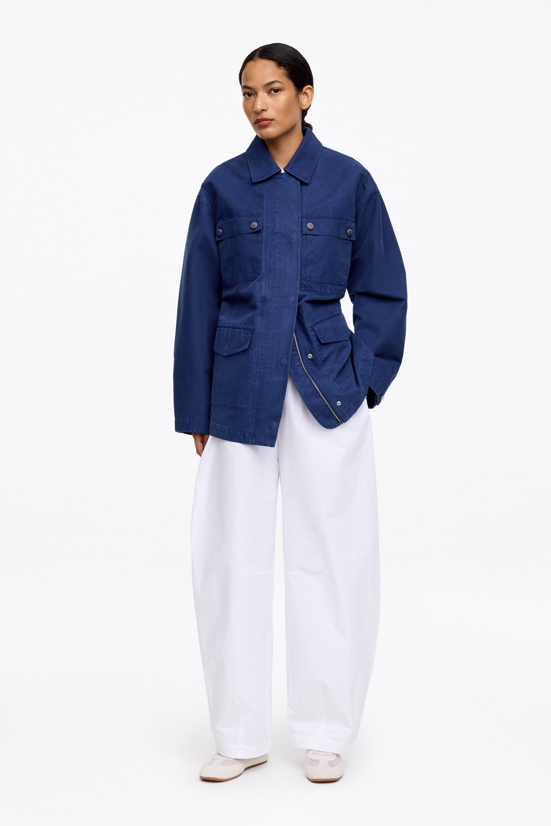 Agrandir l'image: Une veste utilitaire bleu profond est présentée de face, dotée d'un col pointu, de deux poches poitrine à rabat avec fermetures à pression, et de deux poches à rabat plus grandes au niveau des hanches. La veste montre une fermeture avant partiellement ouverte et est portée sur un pantalon blanc ample à jambes larges.