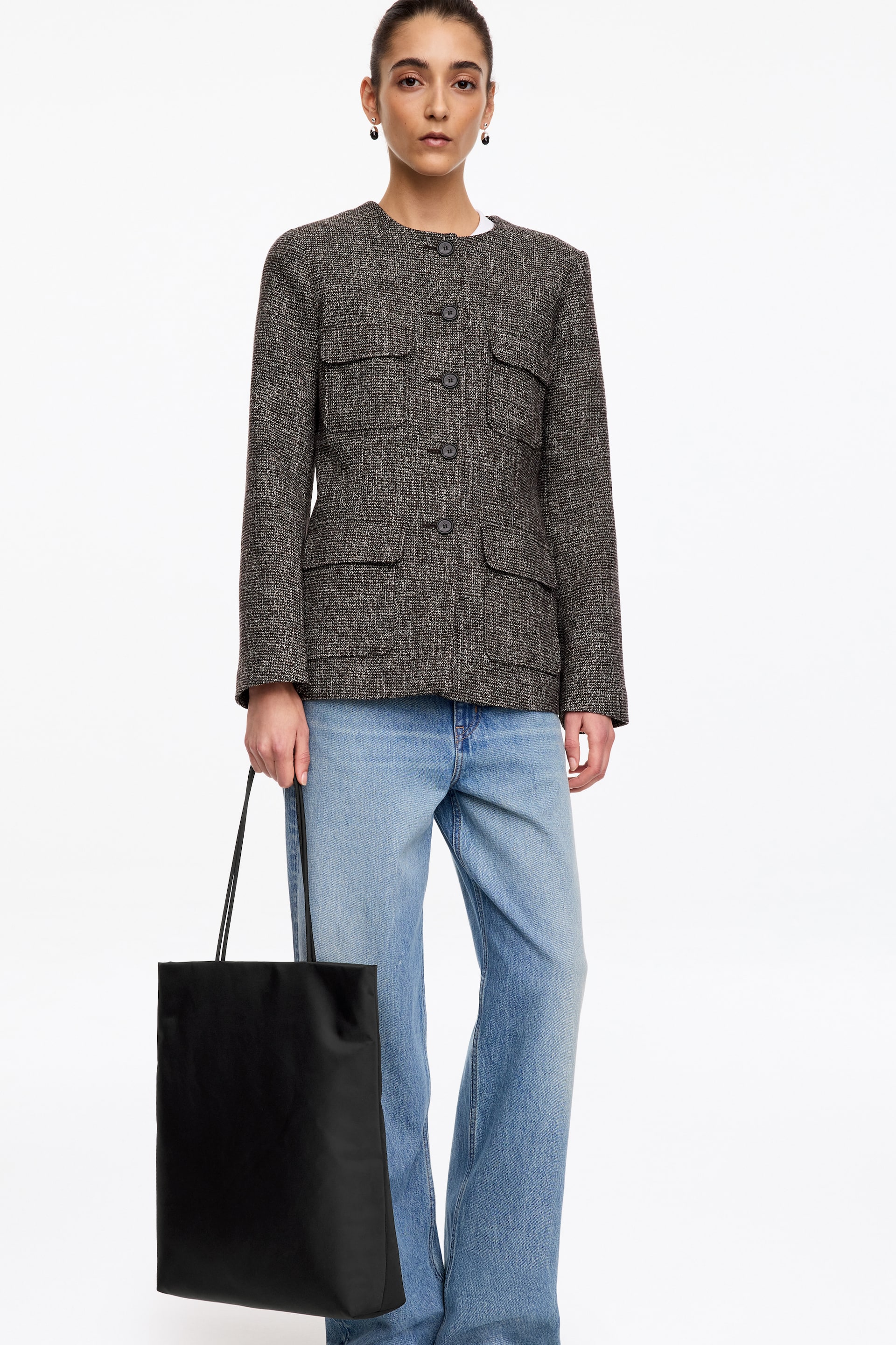 View larger image: A black, rectangular nylon tote bag with thin shoulder straps is being carried by the model, who is holding it by their side. The lightly padded bag is styled with a textured grey jacket and light-blue jeans.