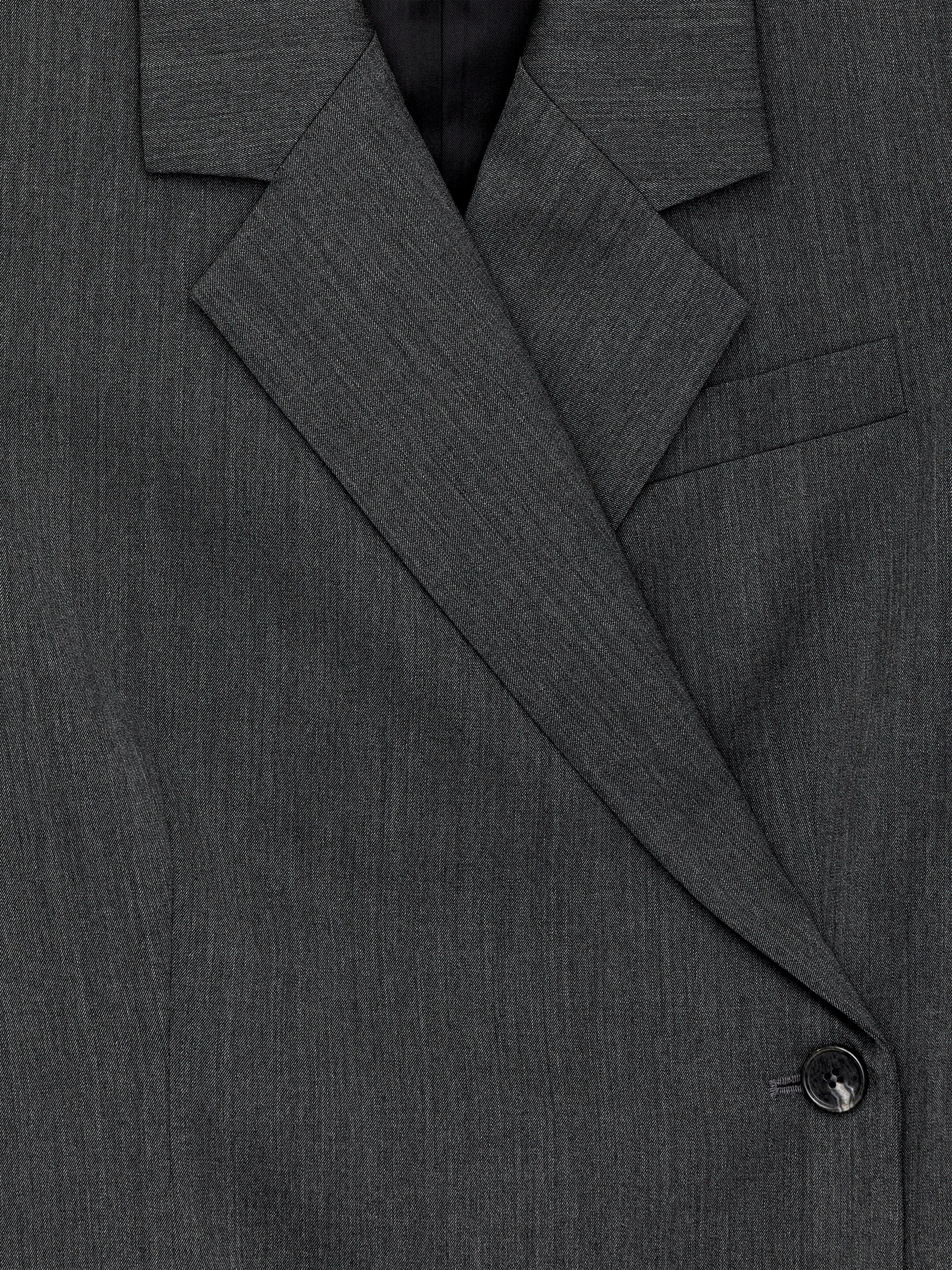 Agrandir l'image: Un blazer croisé ajusté gris foncé est présenté de face, mettant en valeur son large revers angulaire, une doublure sombre visible et une poche passepoilée à rabat unique. Le blazer se ferme avec un bouton foncé, et des coutures de panneaux verticaux sont évidentes.