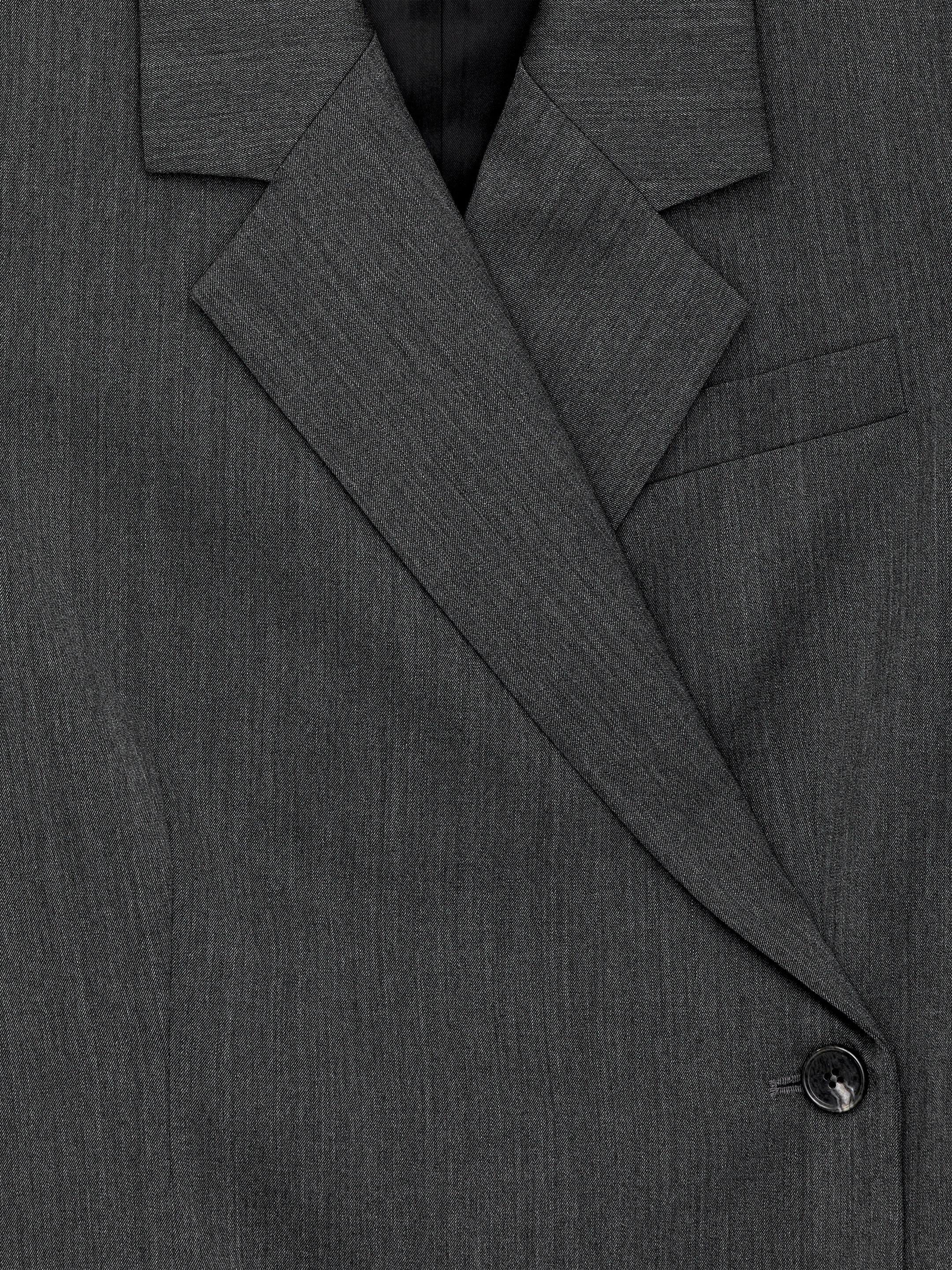 Visualizza immagine più grande: Un blazer avvolgente su misura di colore grigio scuro viene presentato dalla PARTE ANTERIORE, mettendo in mostra il suo ampio revers angolato, una fodera scura visibile e una singola tasca a filetto con patta. Il blazer si chiude con un bottone scuro e sono evidenti le cuciture verticali dei pannelli.