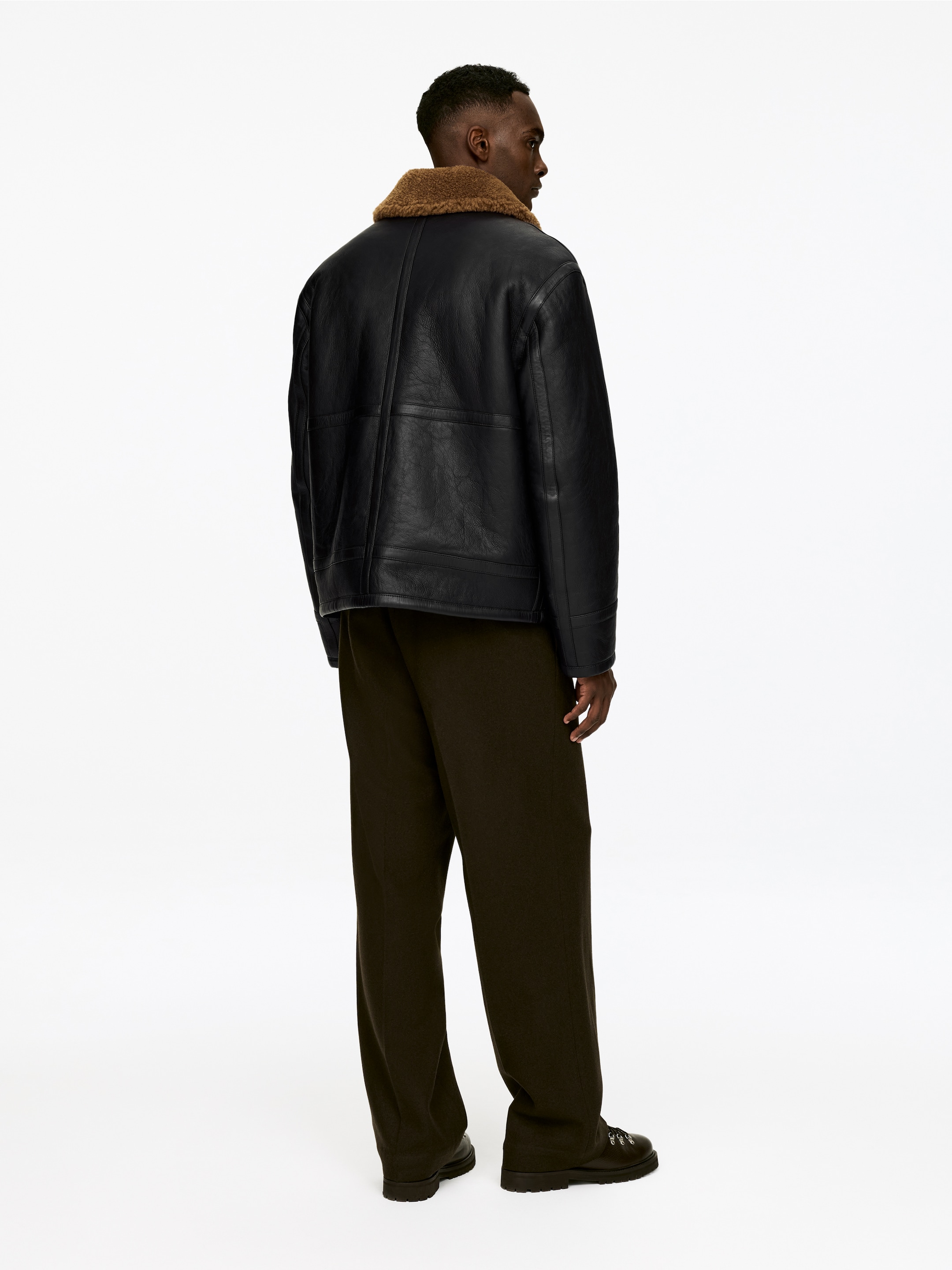 Agrandir l'image: Une veste en simili cuir noir est présentée de l'arrière-droit, avec un col en fausse fourrure marron et des détails de panneaux visibles sur son dos. La veste est associée à un pantalon vert olive à jambes larges et des bottes à semelles épaisses et foncées avec un laçage à anneaux métalliques en D.