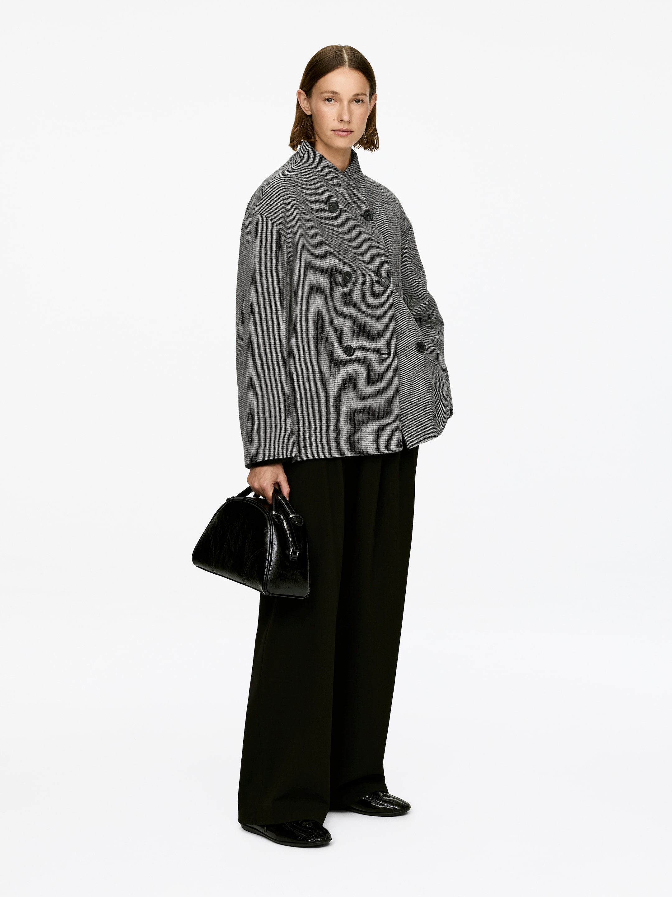 Grösseres Bild ansehen: Eine zweireihige Jacke, gefertigt aus einem grauen Wollmischgewebe mit einem dezenten Muster, wird vom Model getragen. Die Jacke hat eine übergroße Silhouette, einen Stehkragen und dunkle Knopfverschlüsse, wobei eine Hand in einer Seitentasche ruht, während das Model eine schwarze Handtasche trägt.