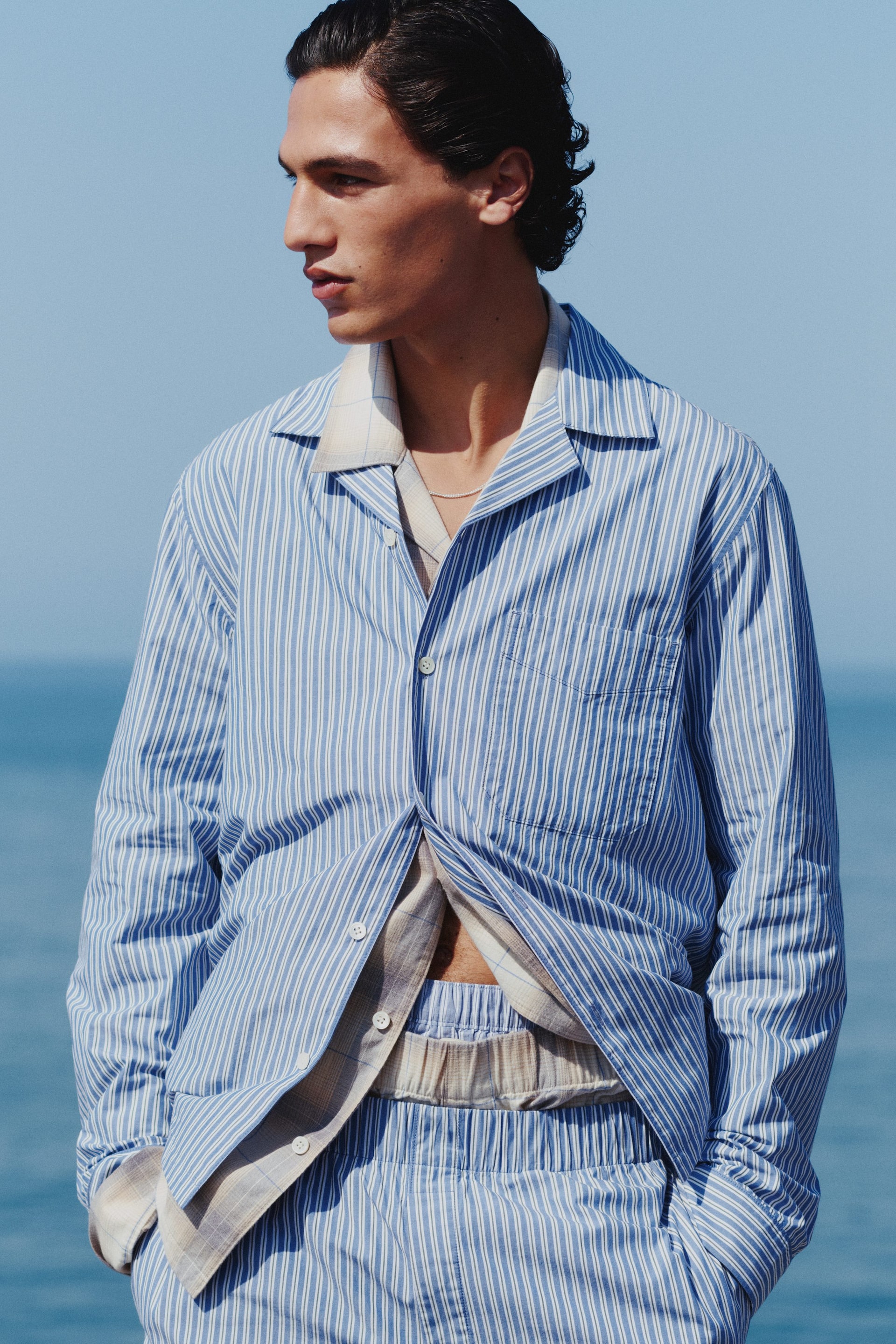 Näytä suurempi kuva: Sinivalkoinen raidallinen pyjamasetti on puettuna, esillä etupuolelta. Pitkähihainen paita, jossa on leirikousi ja vasemman rinnan paikkatasku, on stailattu avoinna kermanvärisen ruutupaidan päällä, ja yhteensopivat raidalliset shortsit joustavalla vyötärönauha täydentävät setin.