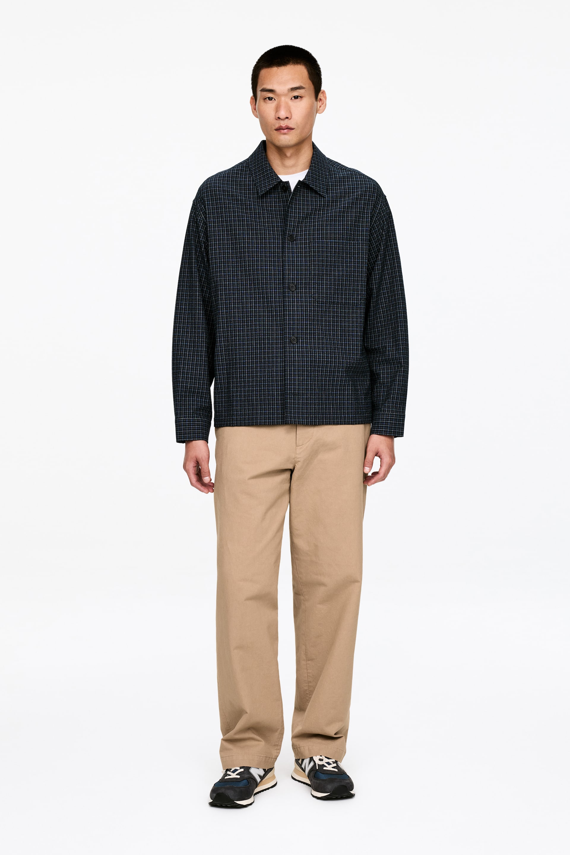 Visa större bild: En mörkblå långärmad skjorta med ett fint rutmönster och en klassisk krage visas framifrån, knäppt över en vit t-shirt. Den har en avslappnad passform med en enkel bröstficka och är kombinerad med ljusbruna vida byxor med en rak, lös skärning.