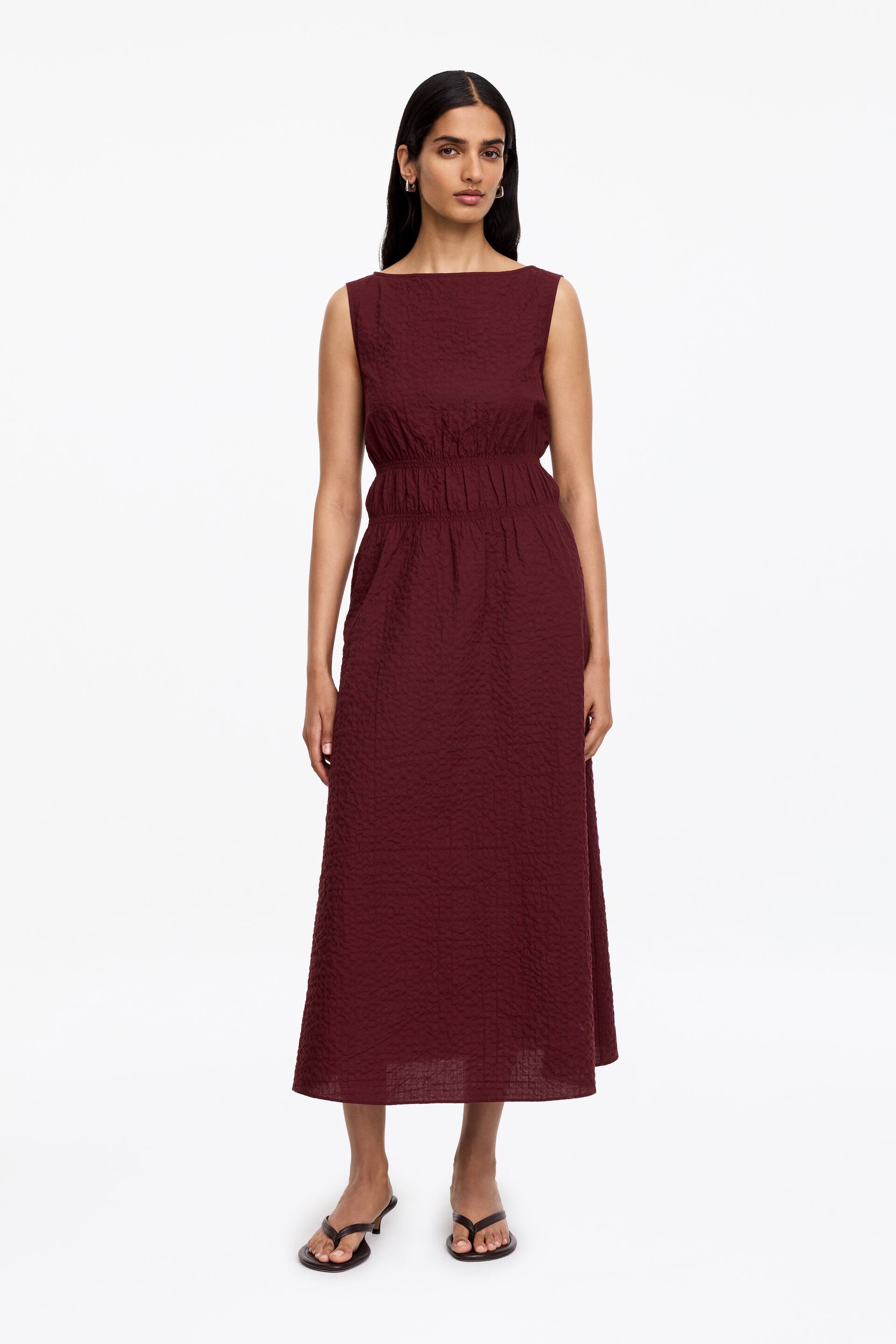 Vis større bilde: Den burgunderrøde ermeløse kjolen er vendt forover, og viser frem en båthals og en krøllete tekstur, med en samlet elastisk midje som flyter ut i et A-linje skjørt som rekker til midt på leggen.