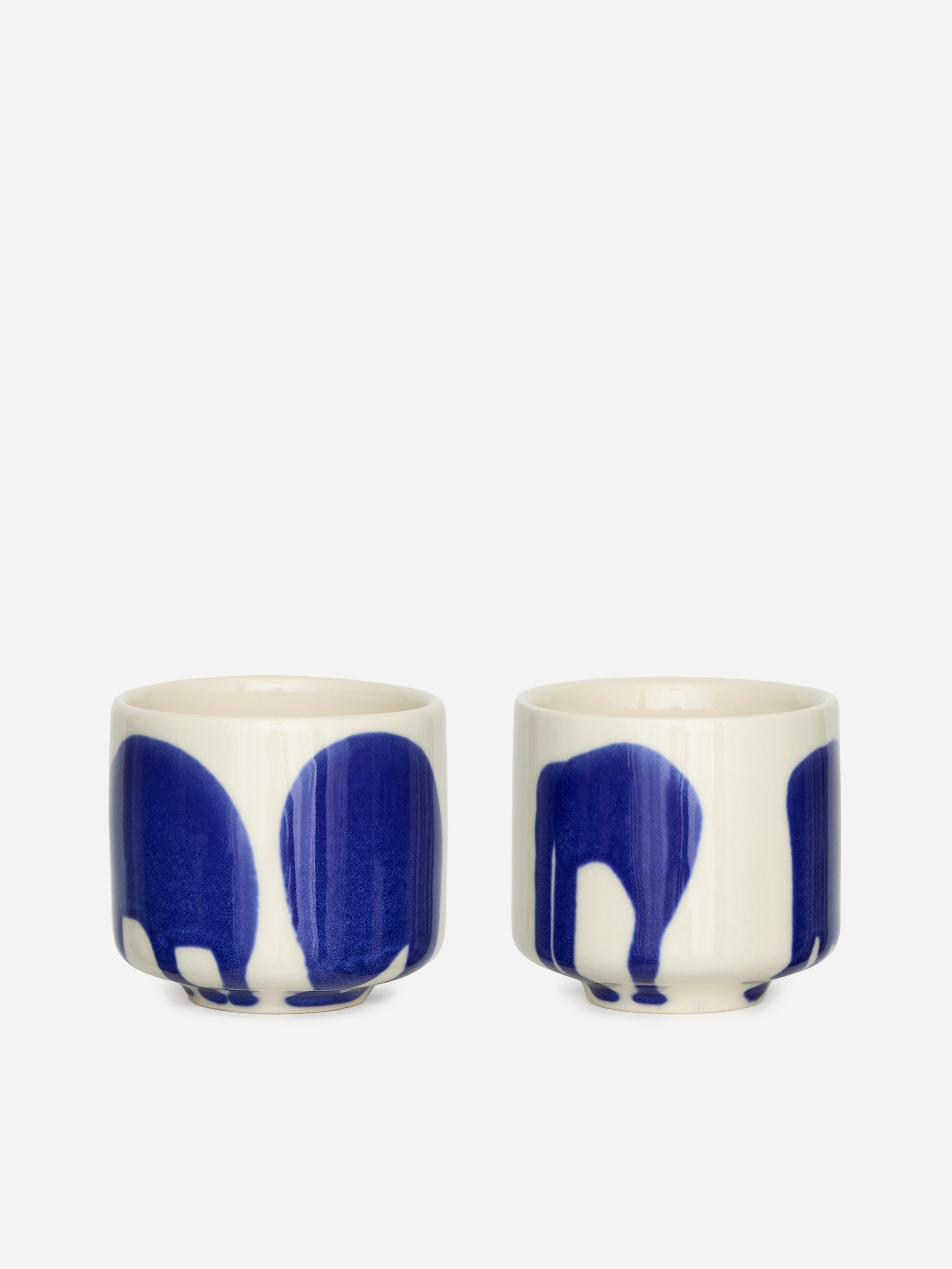 View larger image: Two glazed stoneware cups are presented FRONT, each featuring a cream base with large, irregular dark blue patterns resembling drips extending from the rim, and resting on a slightly raised base.