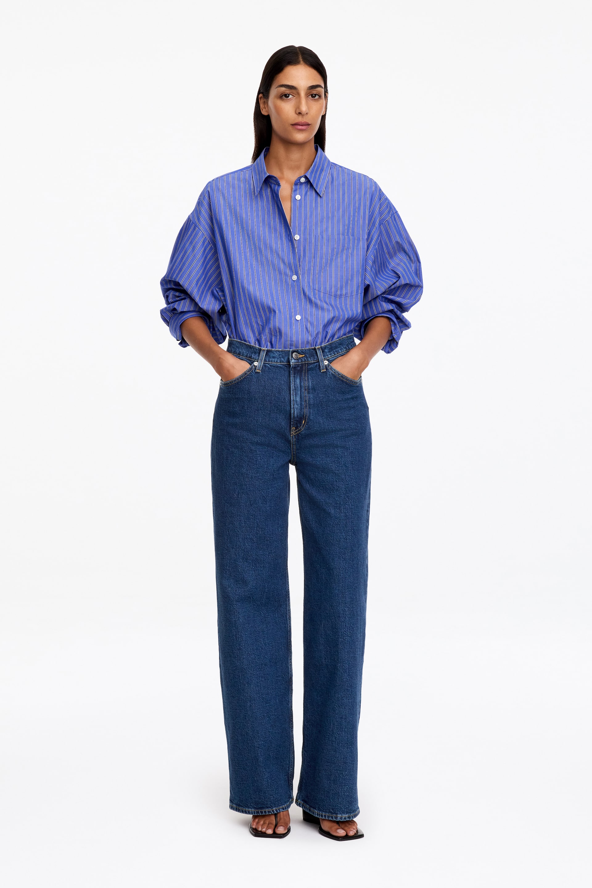 Agrandir l'image: Le mannequin porte un jean en denim bleu foncé taille haute, les mains glissées dans les poches avant. Une chemise boutonnée rayée bleu et blanc est rentrée dans la ceinture, mettant en valeur la coupe taille haute du jean.
